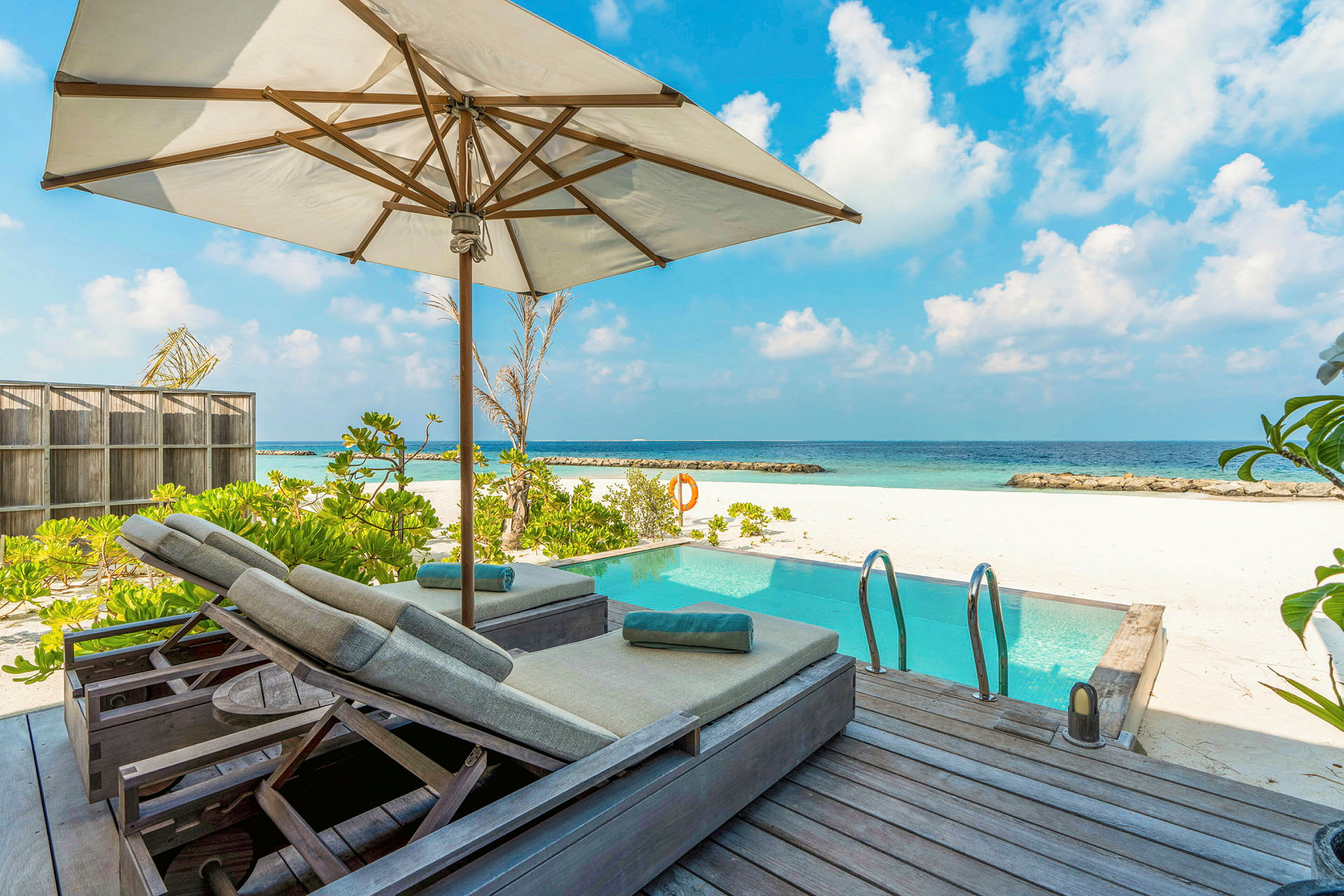 Billede af en solterrasse med parasol og liggestole ved siden af en pool, med hvid sandstrand og hav i baggrunden. (AI-genereret tekst)