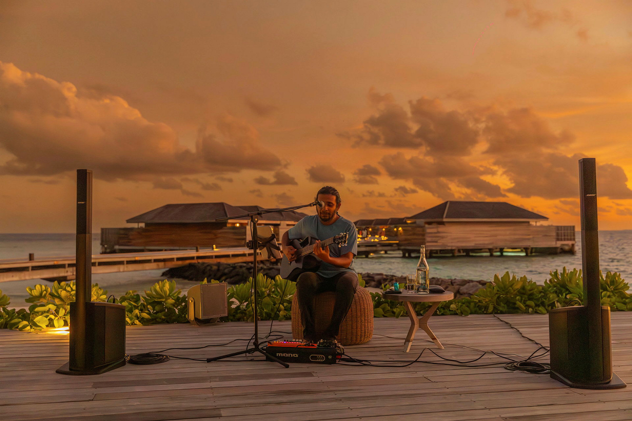 En person spiller guitar, mens solen går ned ved en strand med vandbungalows. (AI-genereret tekst)