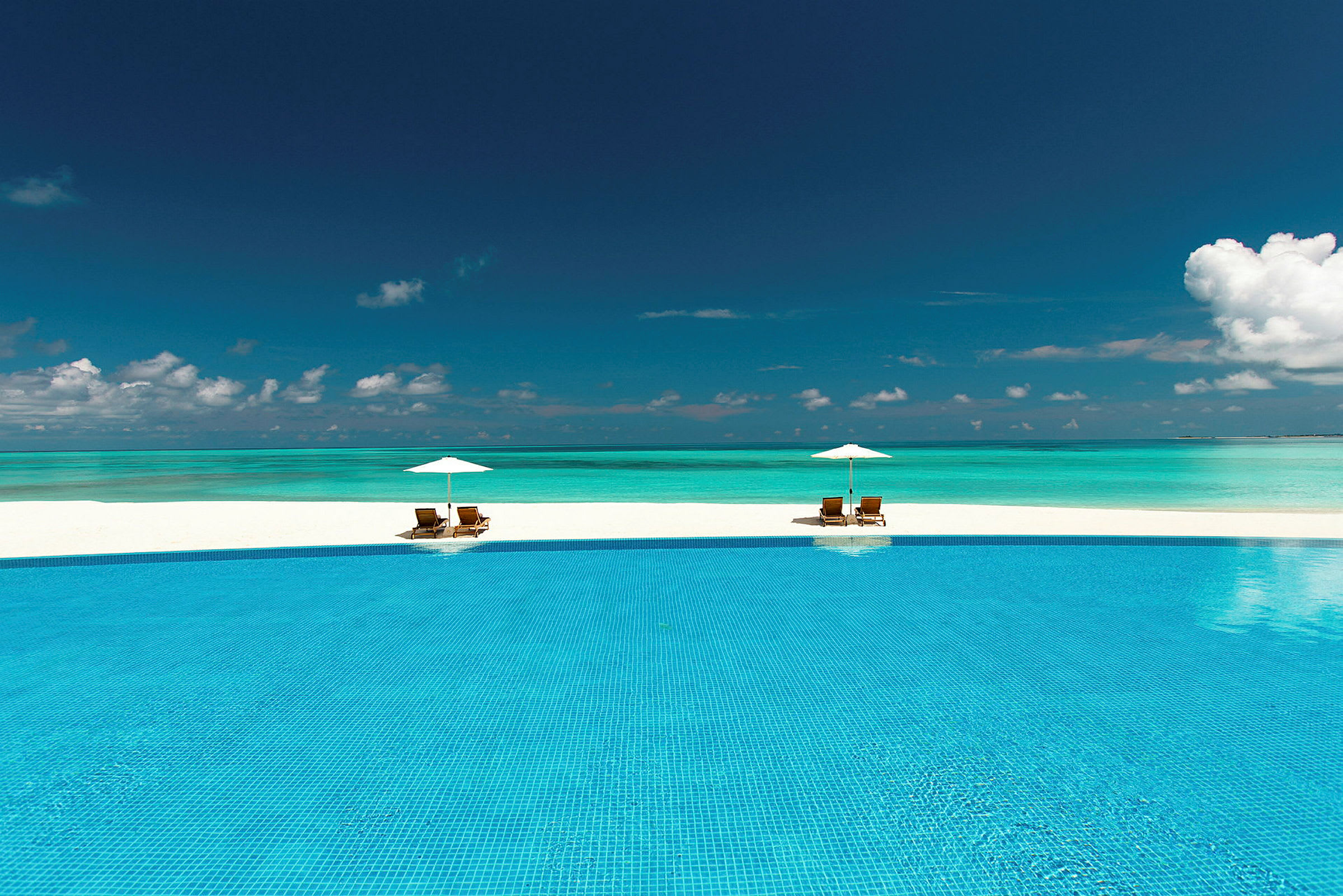 En infinitypool foran en paradisstrand med to liggestole og parasoller. (AI-genereret tekst)