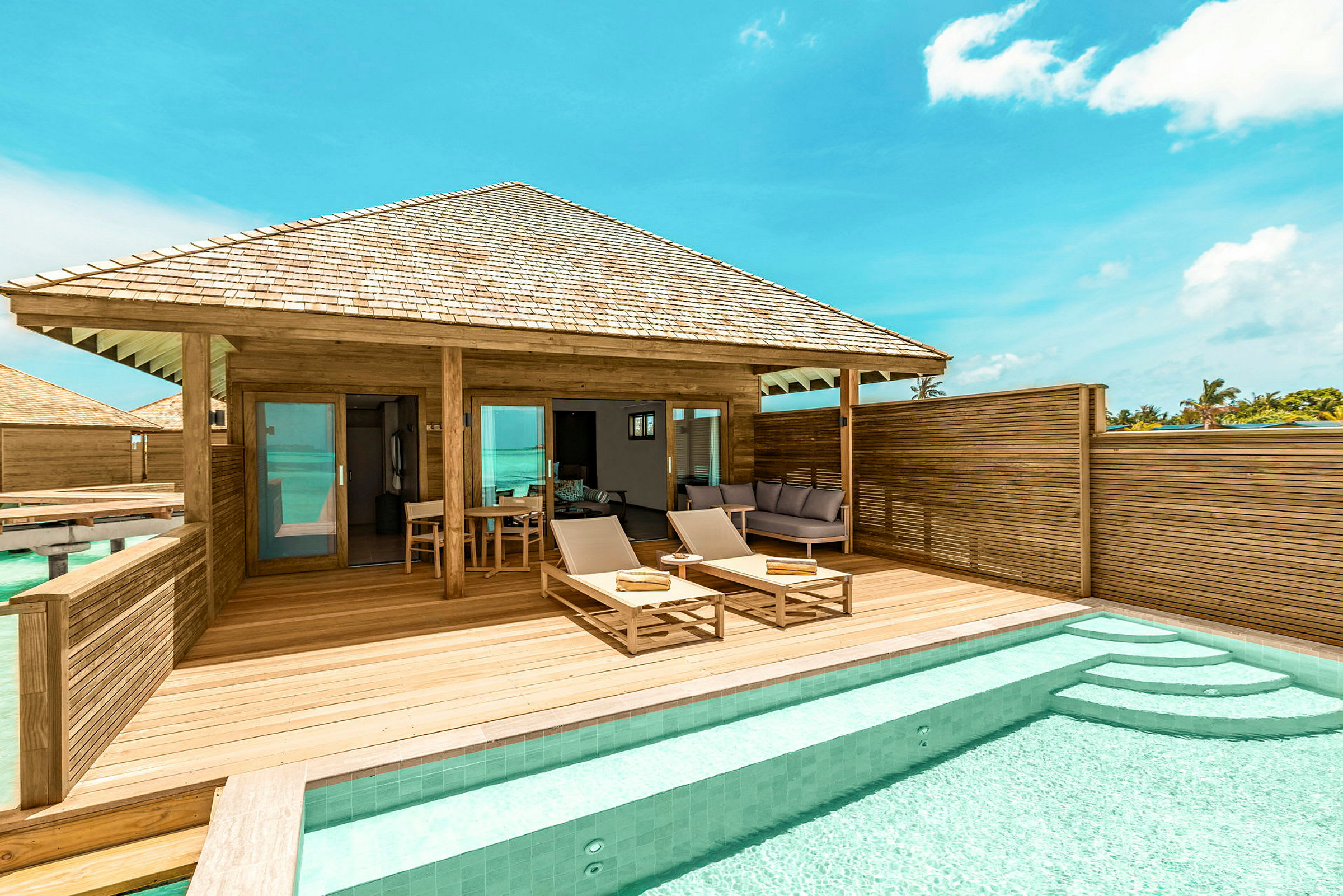 En terrasse med to liggestole, et bord og en privat pool foran en træbungalow. Solrigt vejr og klar blå himmel. (AI-genereret tekst)