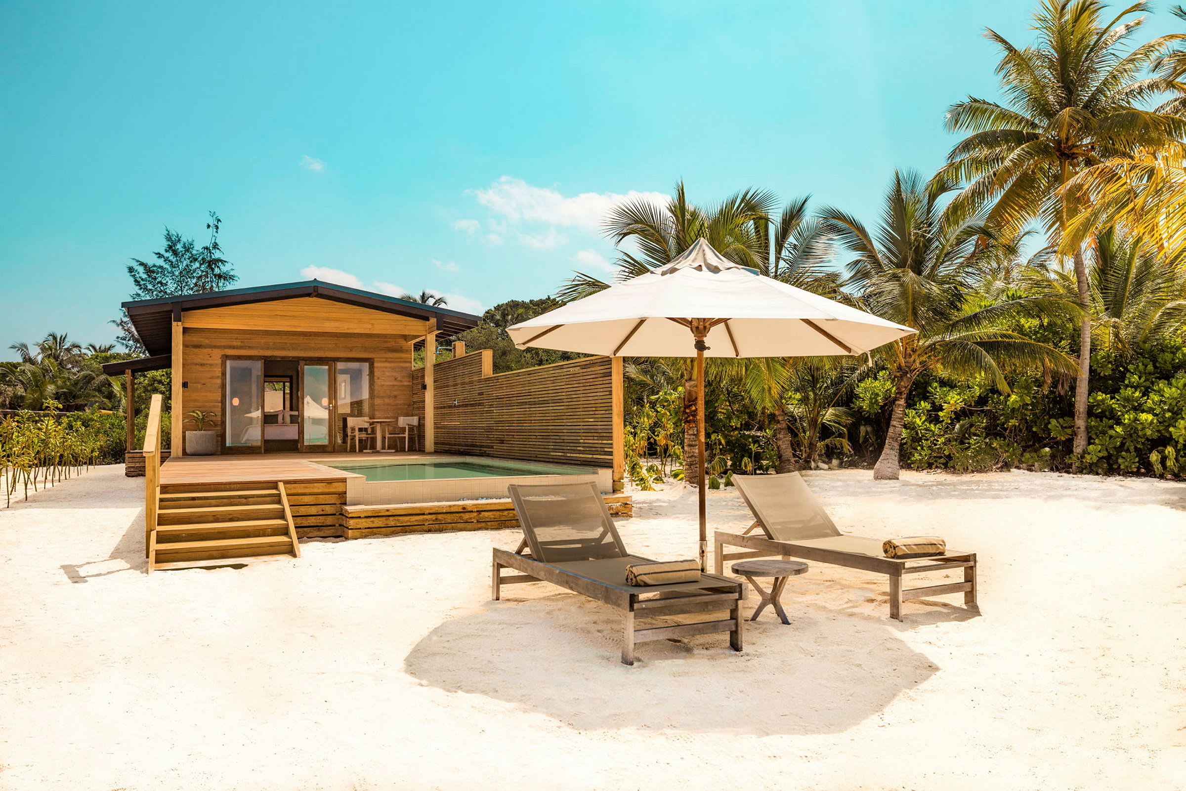 Et sommerhus foran en pool med to solsenge og en parasol på en sandstrand. (AI-genereret tekst)