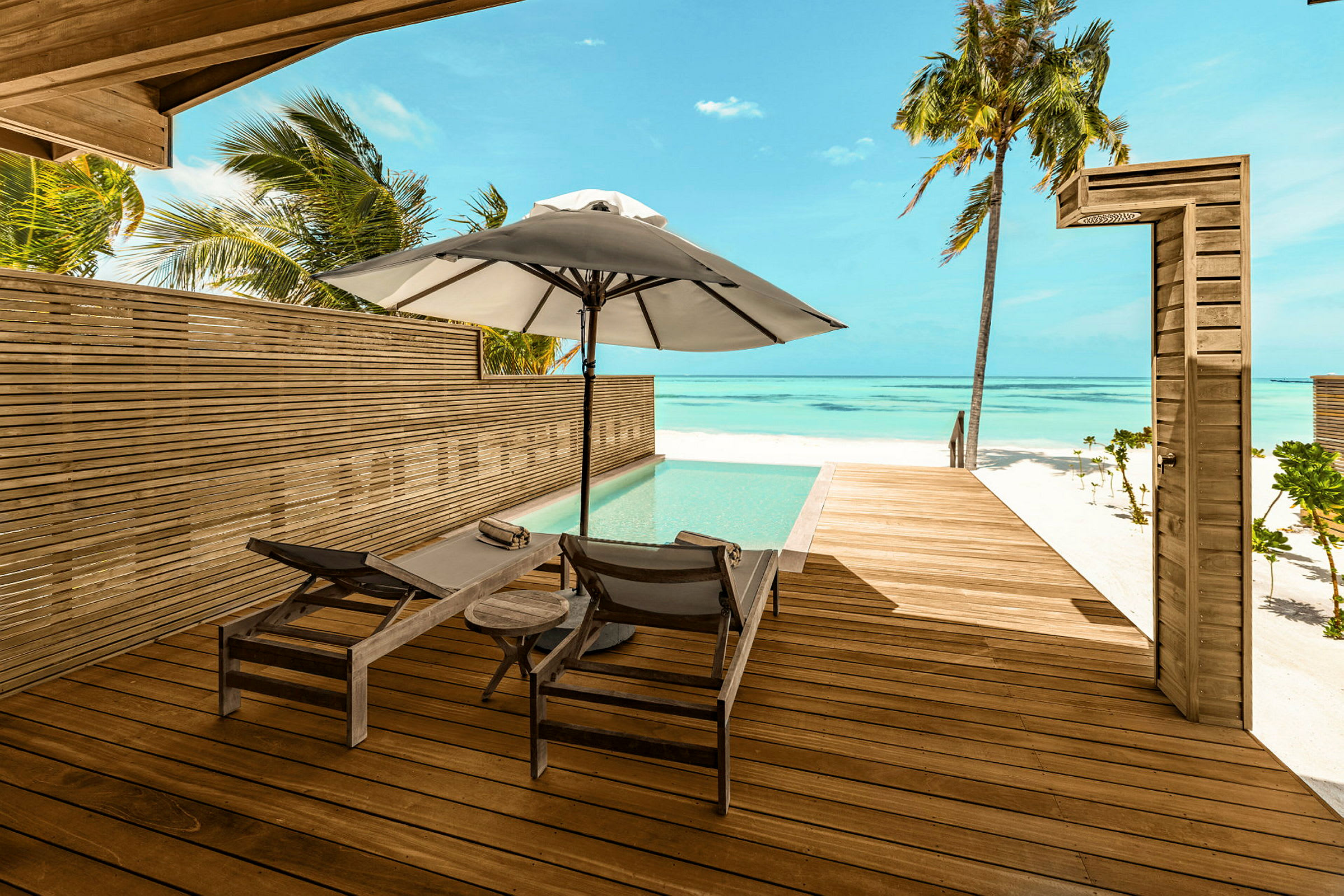 En træterrasse ved en pool med udsigt over stranden og havet. To solsenge og en parasol står på terrassen. (AI-genereret tekst)