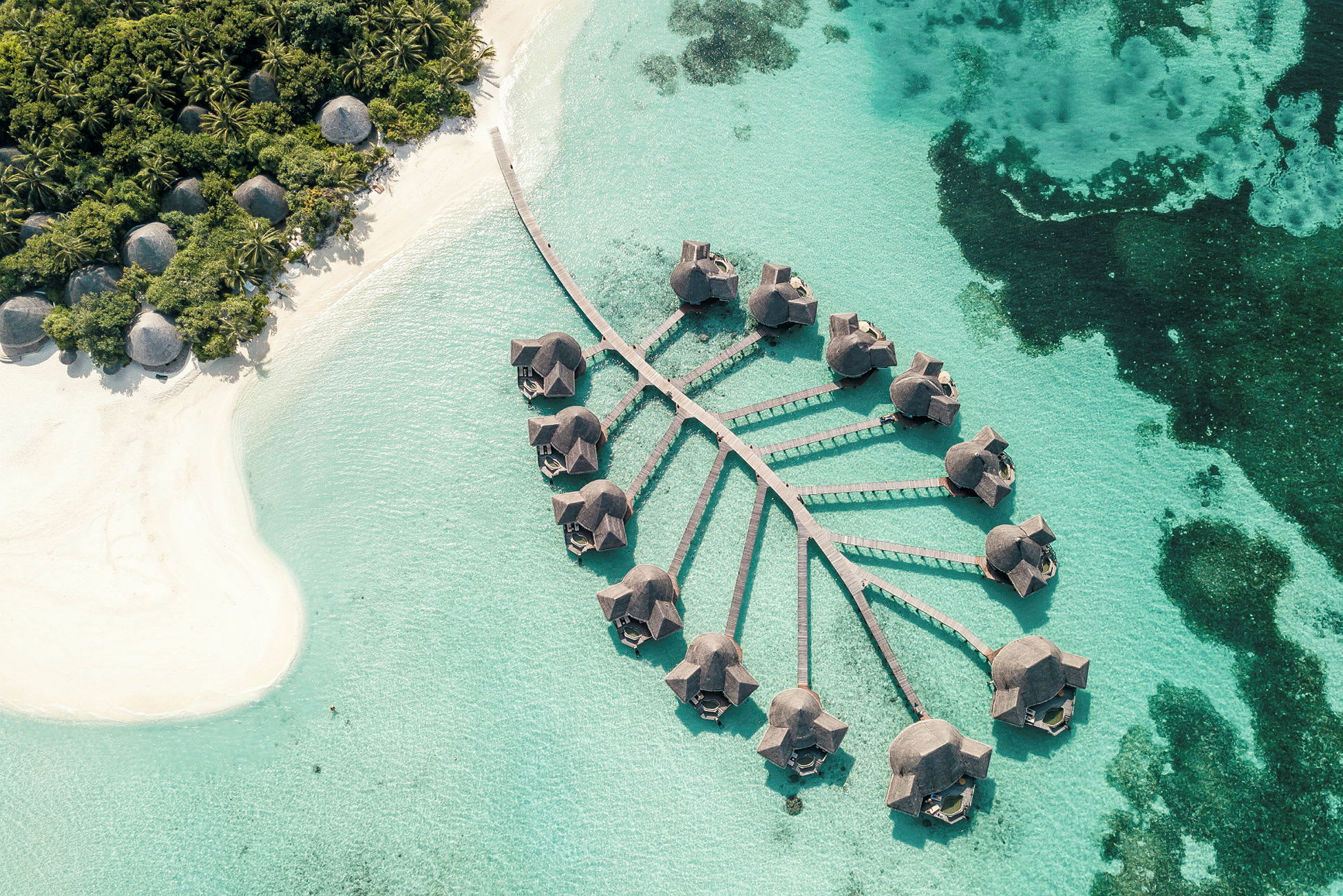 Luftfoto af bungalower på pæle over klart blåt vand med en nærliggende sandstrand og frodig vegetation. (AI-genereret tekst)