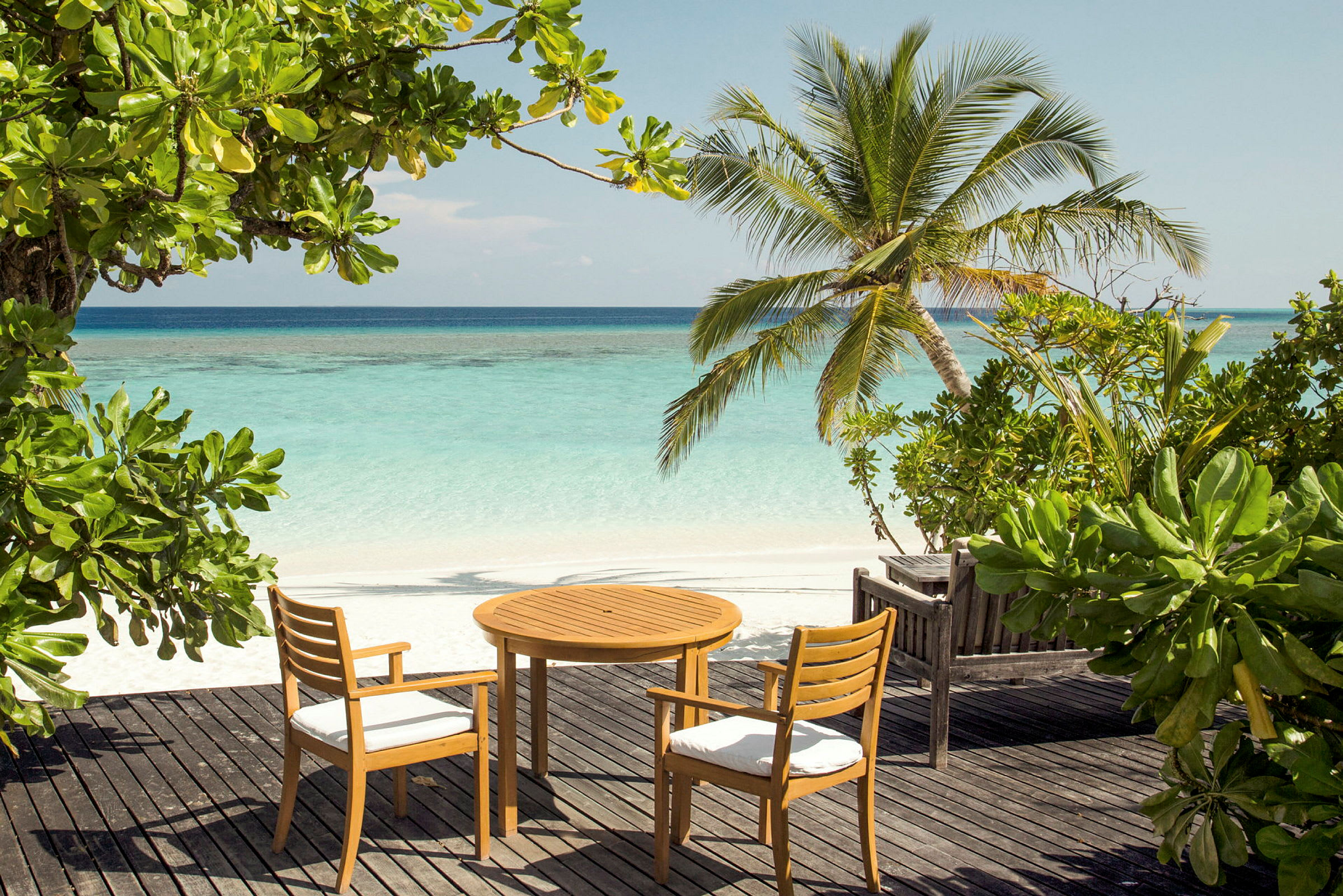 Et træbord og to stole står på en terrasse ved stranden med palmer og udsigt over havet. (AI-genereret tekst)