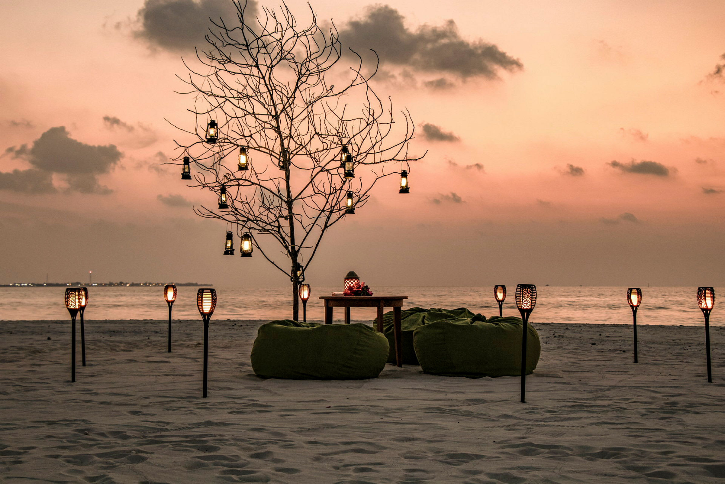 En strandscene ved solnedgang med et dekoreret træ, sækkestole og bord samt aflange lanterner. (AI-genereret tekst)