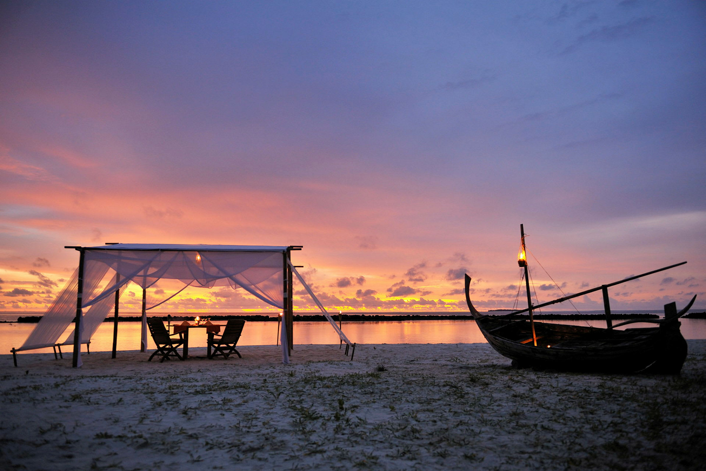 En strand med en lille baldakin og stole ved solnedgang, med en træbåd og lygter. (AI-genereret tekst)