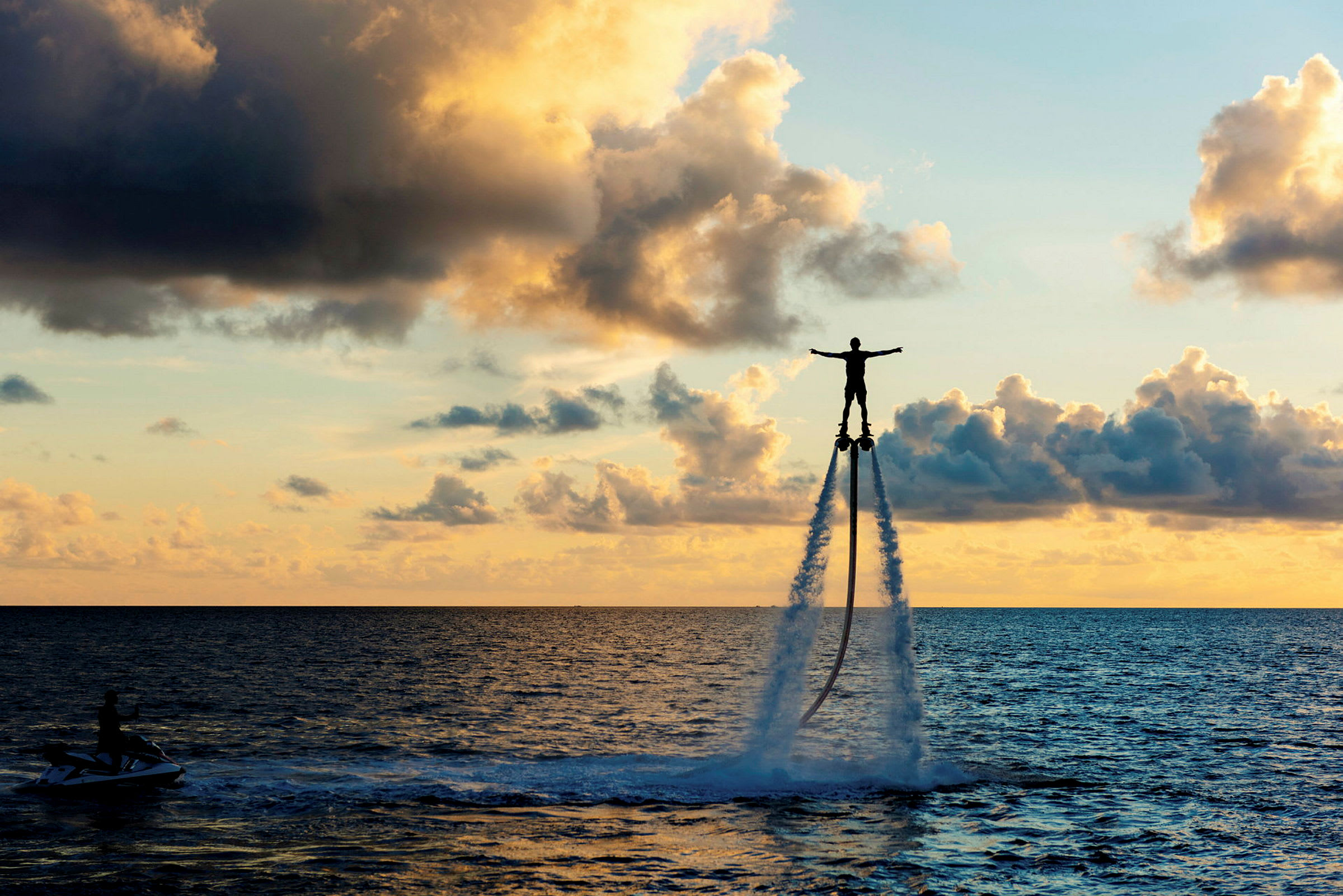 En person flyver over havet ved hjælp af en vandjetpack, mens solen går ned. (AI-genereret tekst)