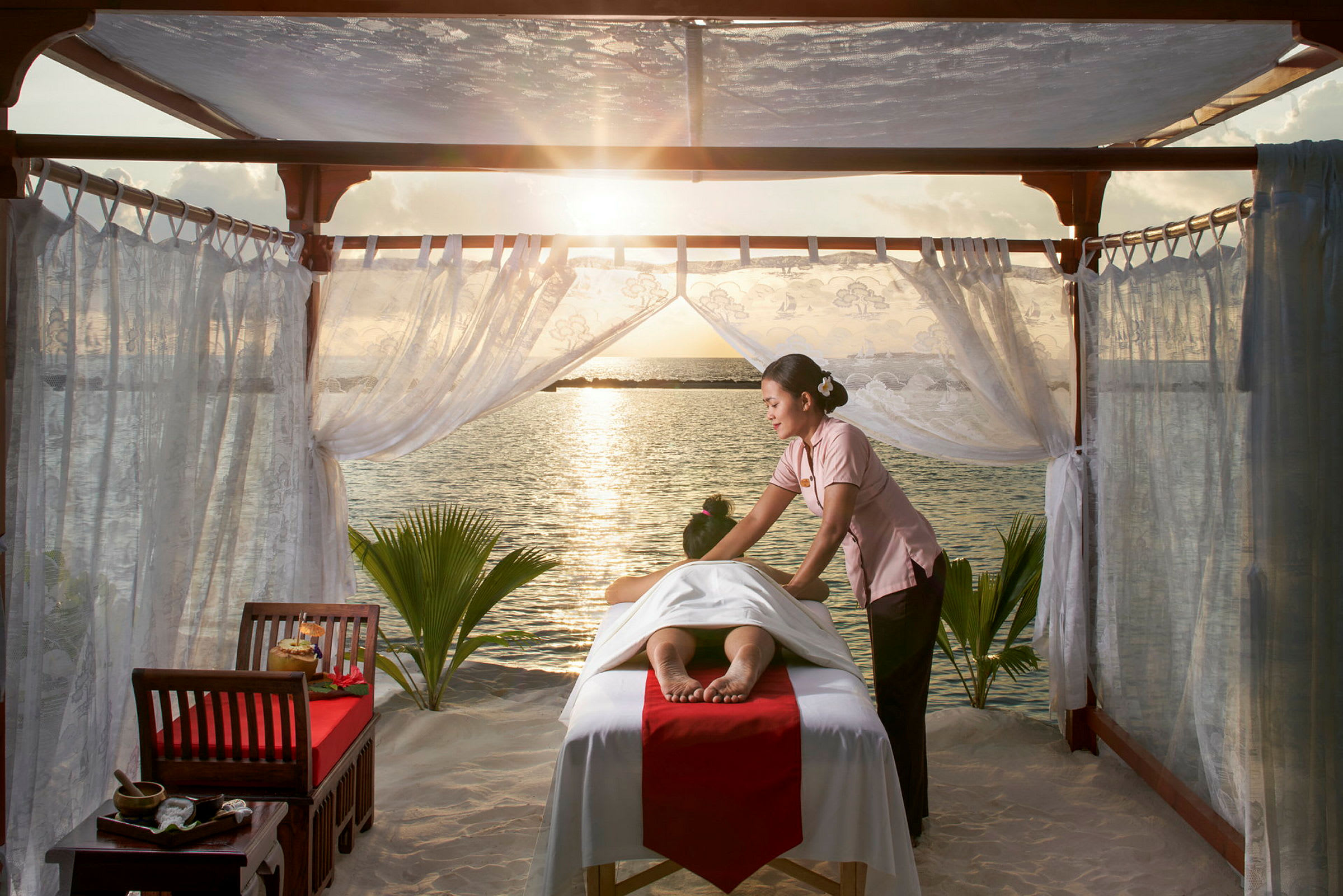 En person modtager rygmassage på en seng under en pavillon ved havet ved solnedgang. (AI-genereret tekst)