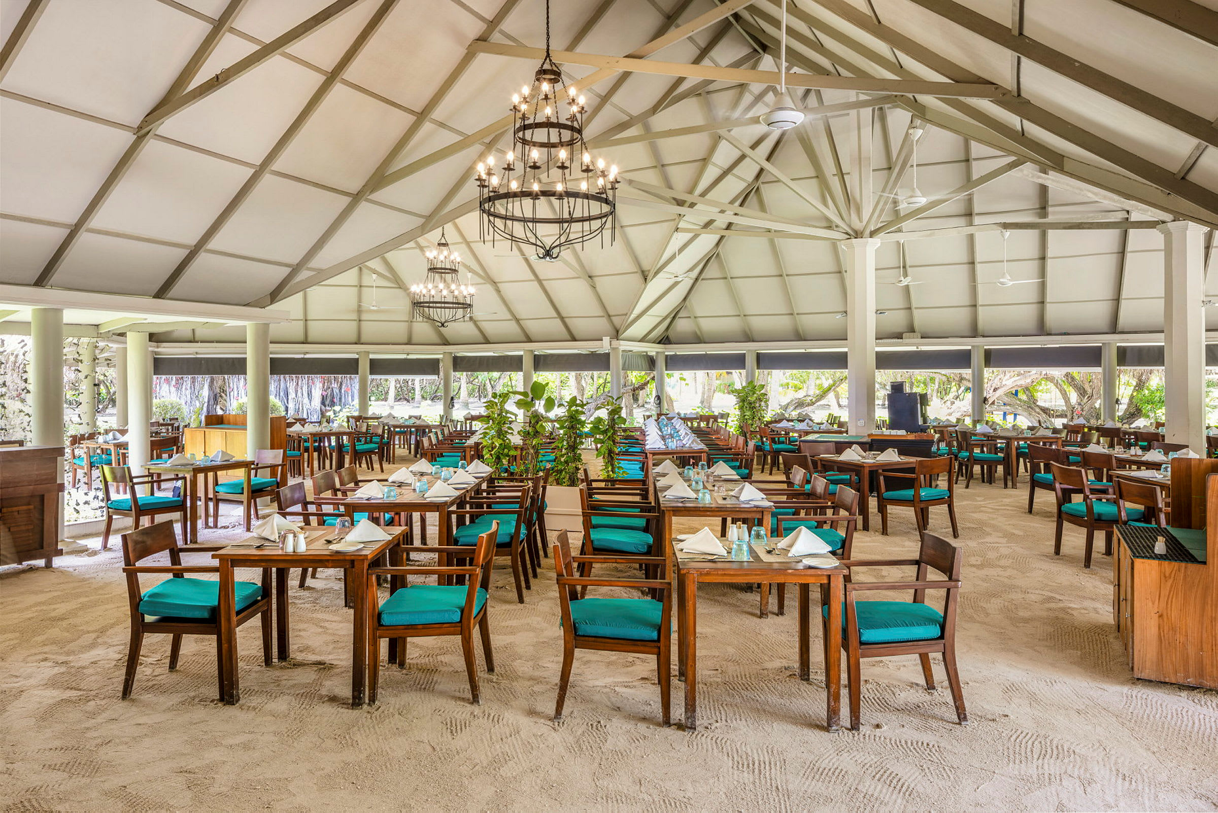 En rummelig restaurant med sandgulv og borde med træstole beklædt med turkise puder under et højt loft med bjælker. (AI-genereret tekst)