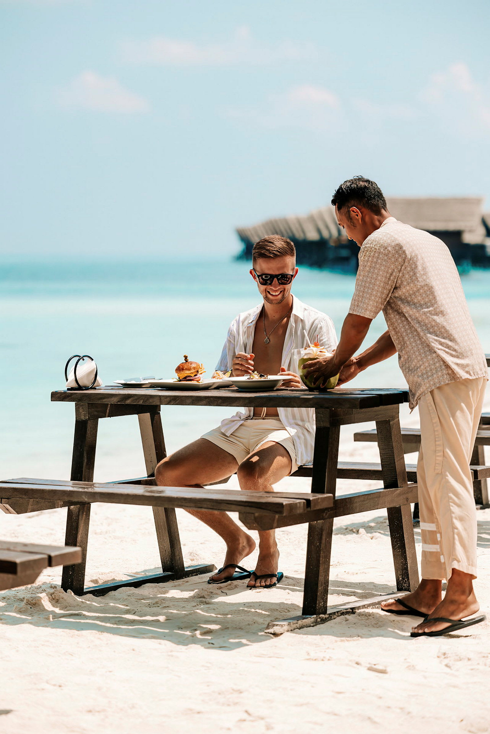 En person sidder ved et bord på en strand med mad og drikke. En anden person serverer og stiller en drink frem. (AI-genereret tekst)