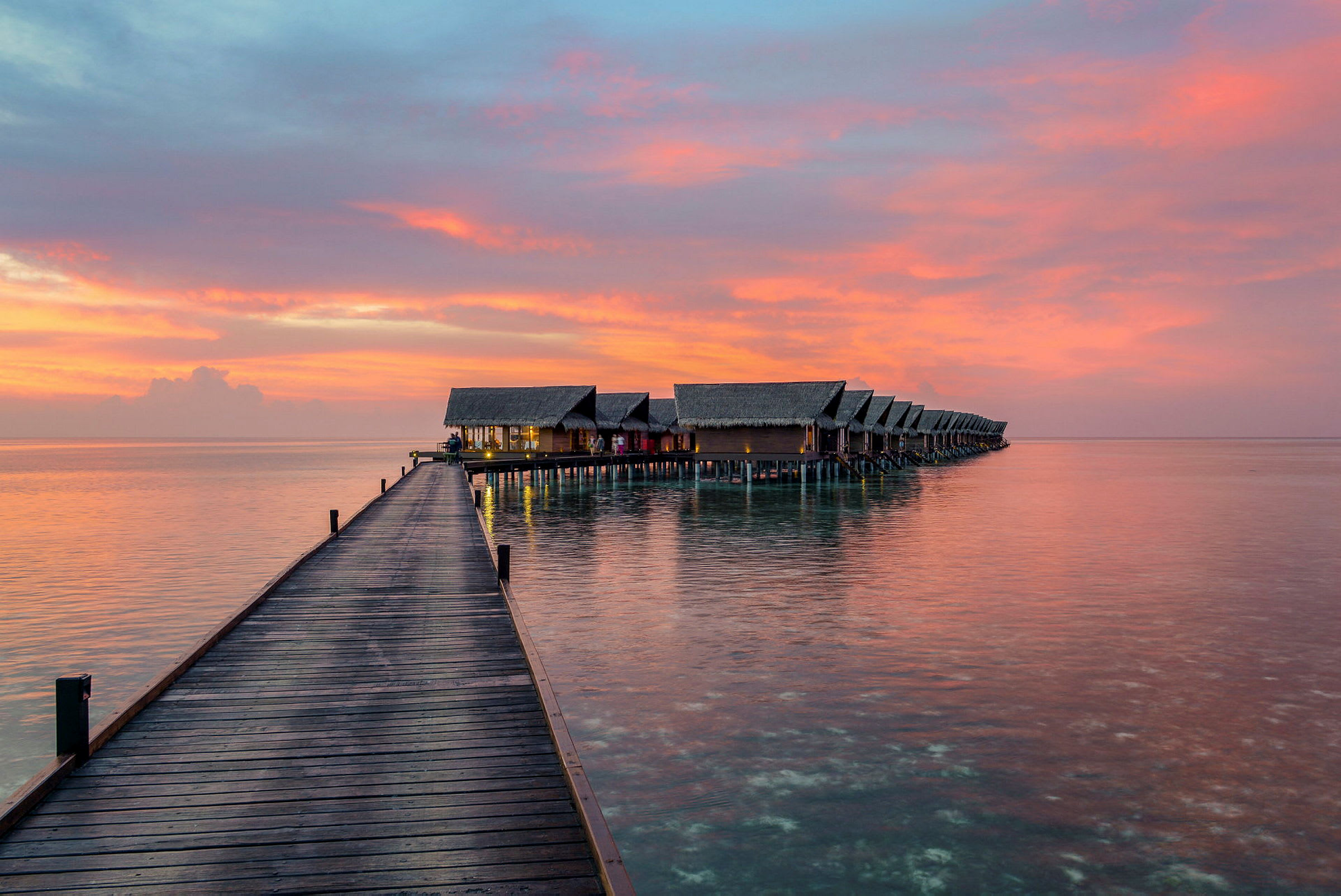 En træbro fører ud til en række overnatningsbungalower ved solnedgang. (AI-genereret tekst)