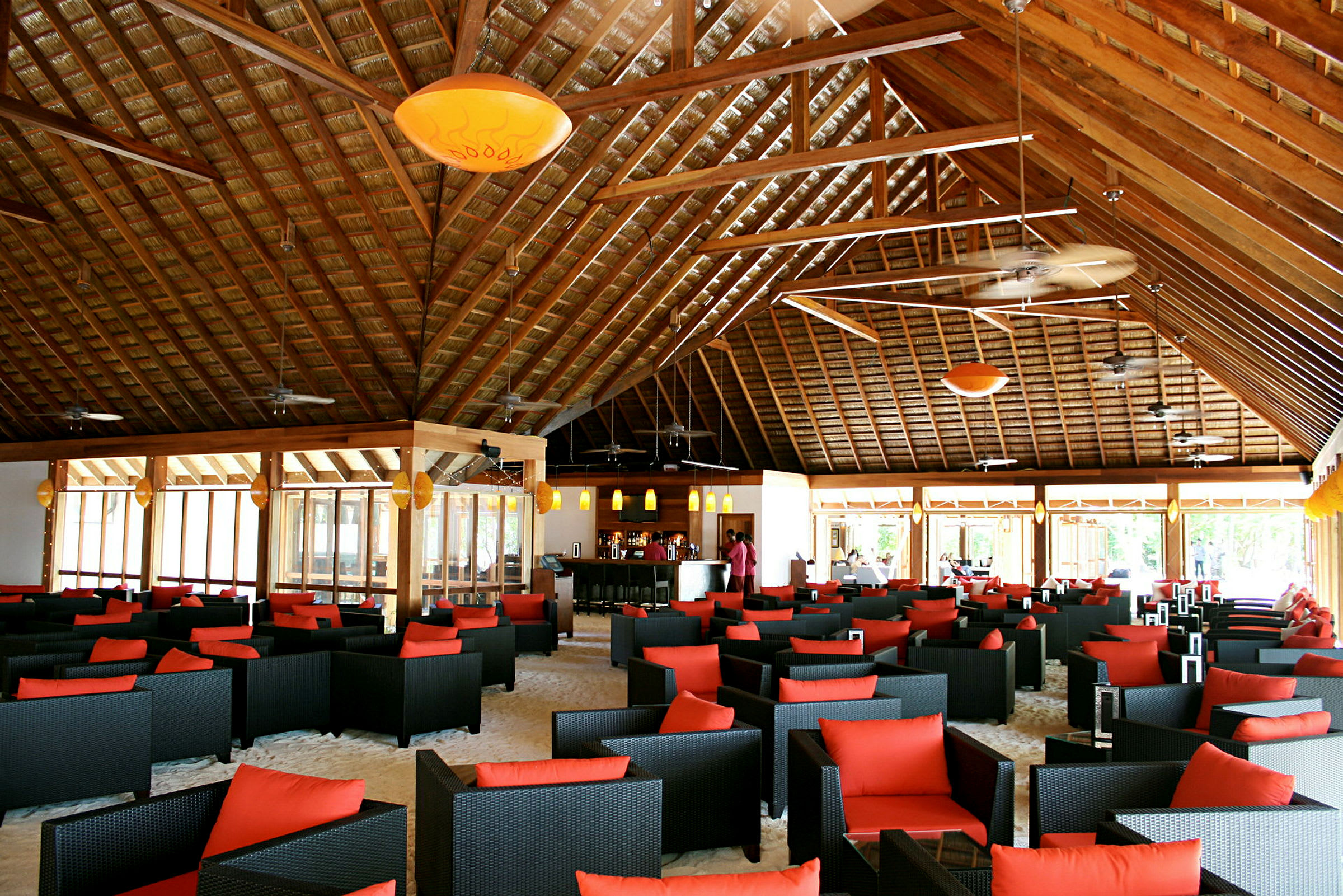 En interiør af en rummelig restaurant eller lounge med sorte flettede stole med røde puder, borde placeret på sandgulv under et højt træbjælkeloft. (AI-genereret tekst)