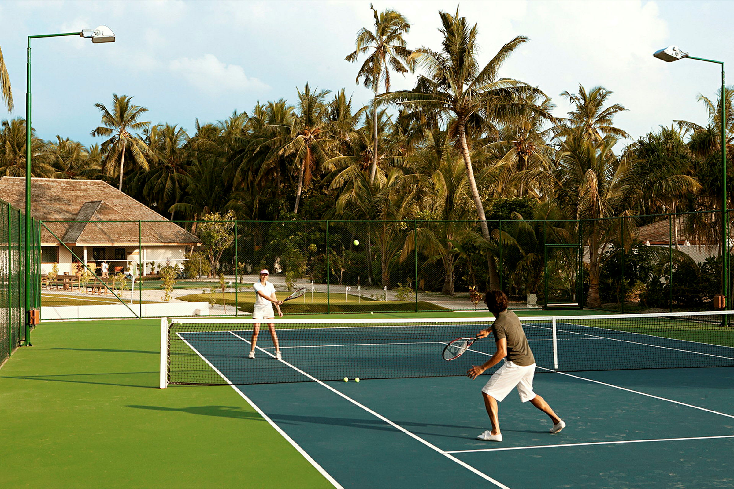To personer spiller tennis på en udendørsbane omgivet af palmer. (AI-genereret tekst)