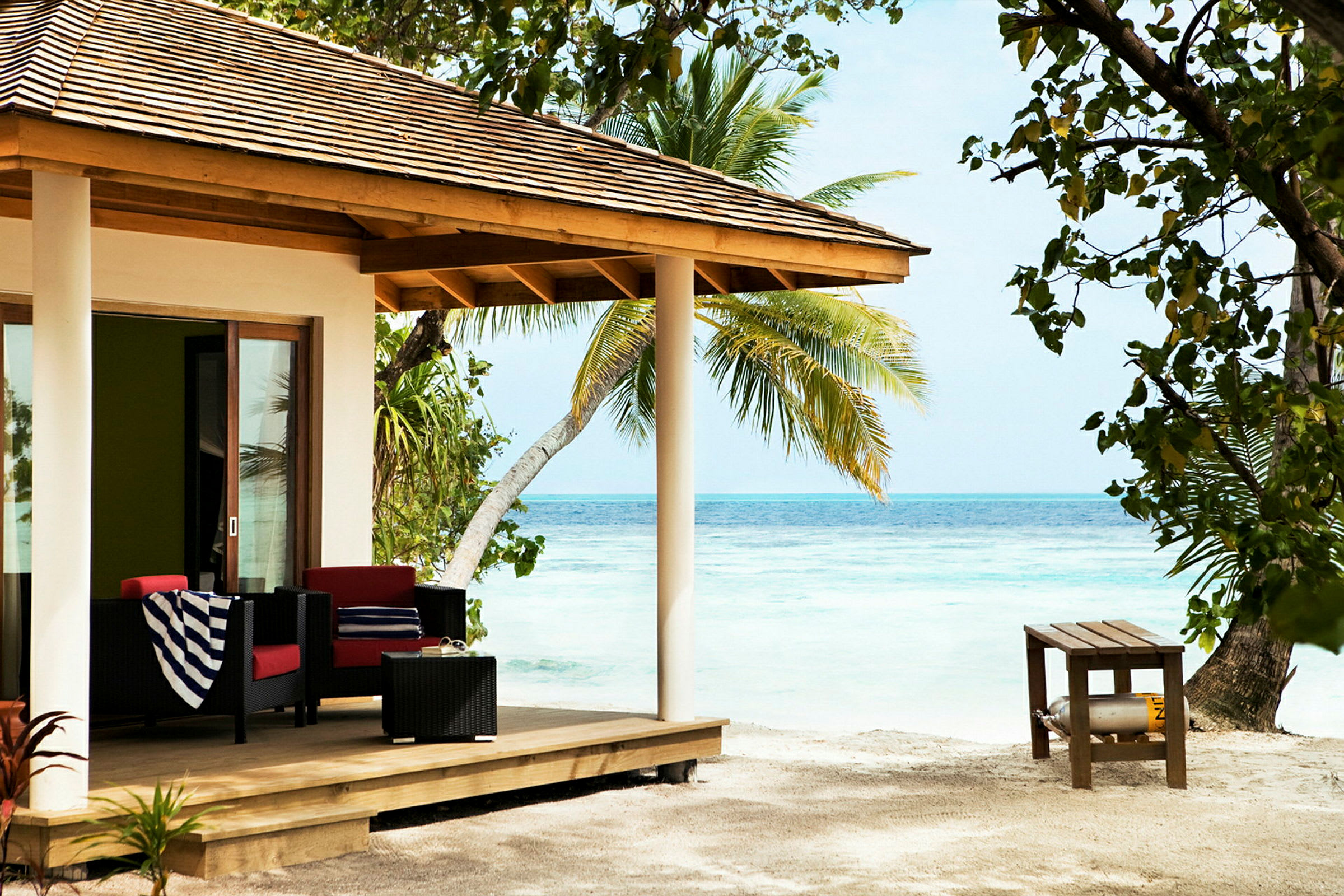 Strandvilla med veranda møbleret med sofaer, bord og dekoreret med palmer omkring. (AI-genereret tekst)