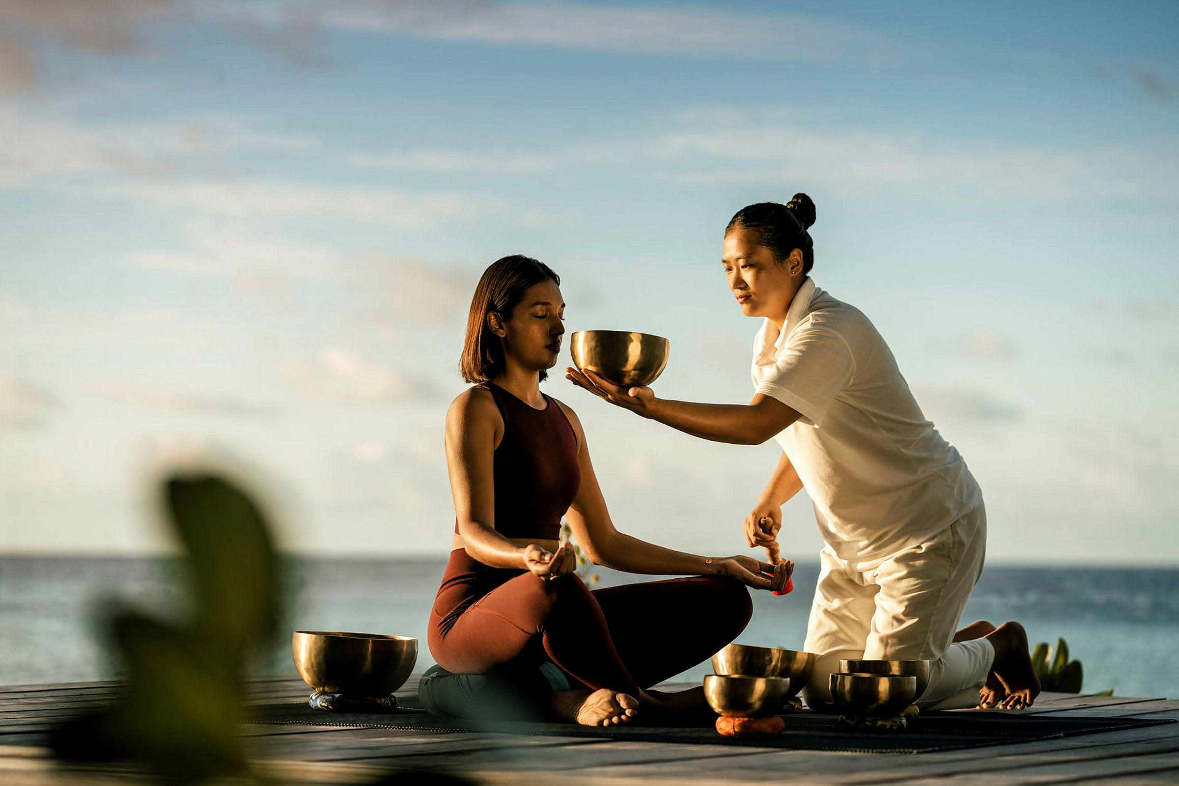 To personer deltager i en meditationssession nær vandet. En holder en klangskål, og den anden sidder med korslagte ben. (AI-genereret tekst)