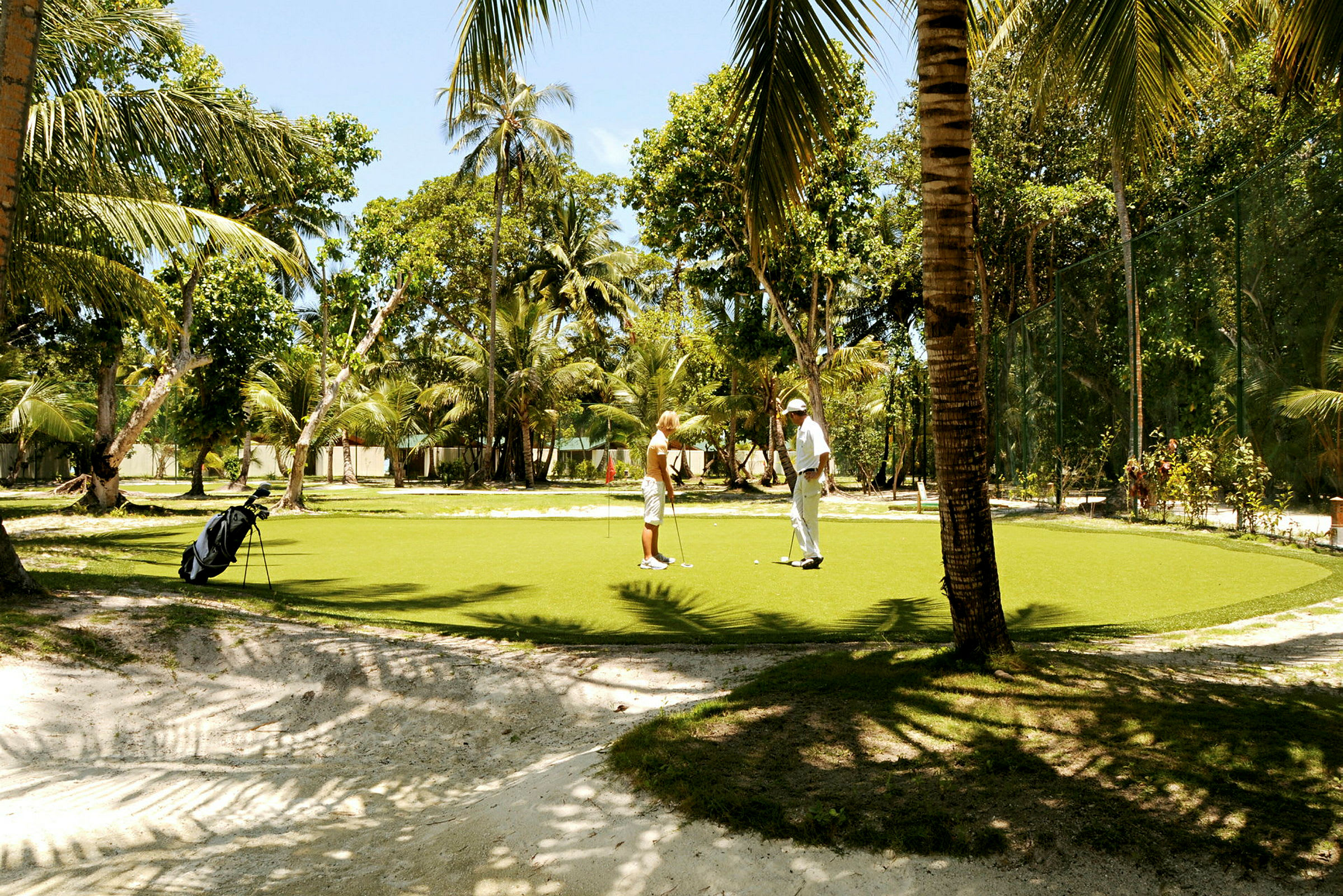 To personer spiller golf på en grøn puttinggreen omgivet af palmer. (AI-genereret tekst)
