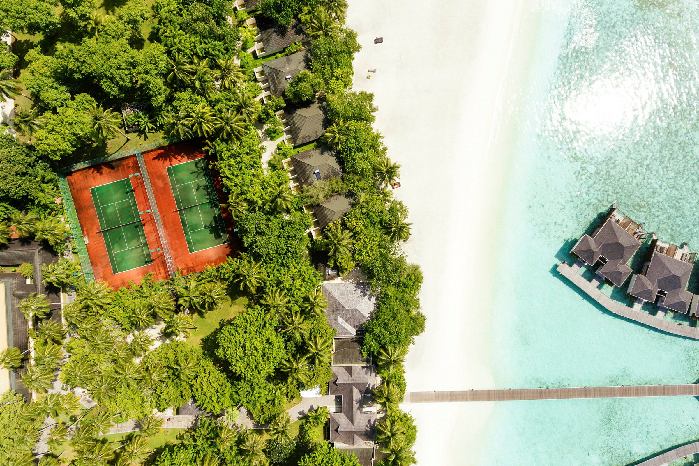 Luftfoto af et tropisk resort ved stranden med tennisbaner, villaer blandt træer og overvandshuse. (AI-genereret tekst)