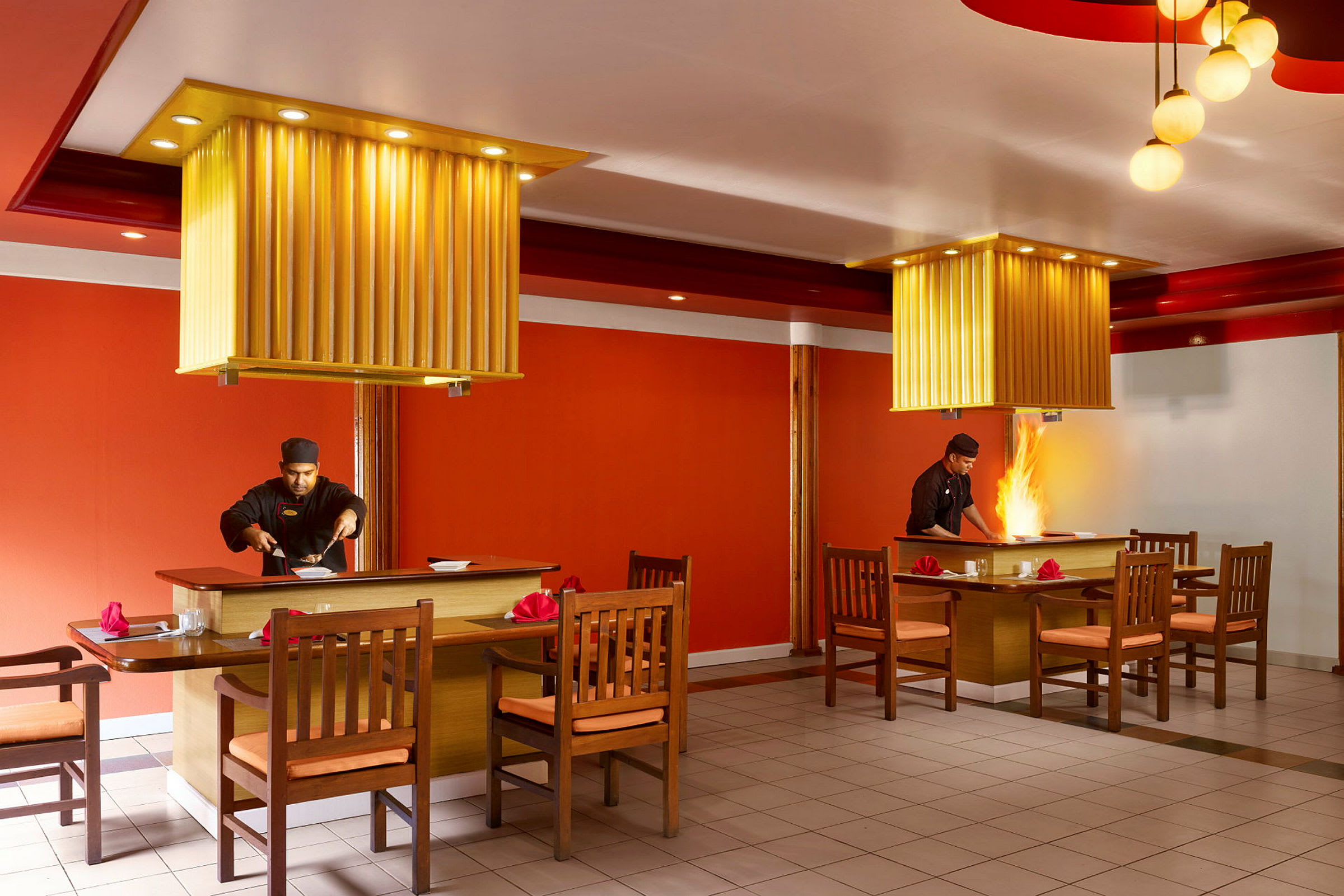 To spiseborde med tilhørende stole i en restaurant med orange interiør og loftslamper. (AI-genereret tekst)