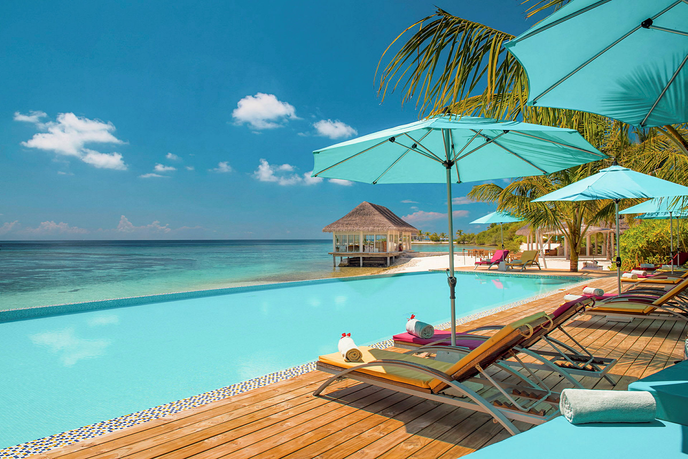 Pool med liggestole og parasoller ved havet, med en strand og en bungalow i baggrunden. (AI-genereret tekst)