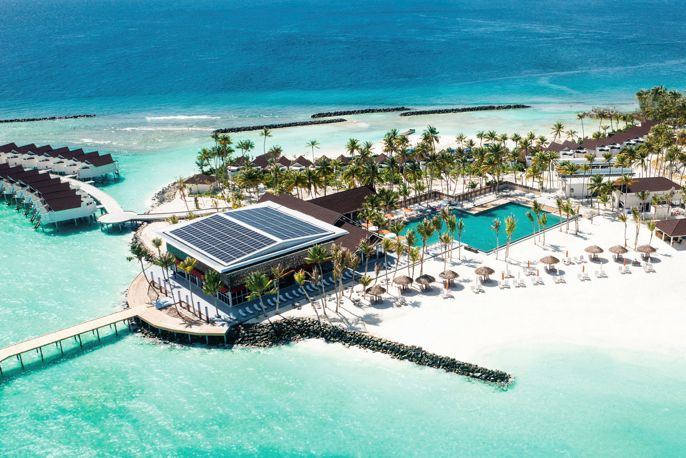 Luftfoto af en resort ved stranden med pool, liggestole og bungalows på vandet. (AI-genereret tekst)