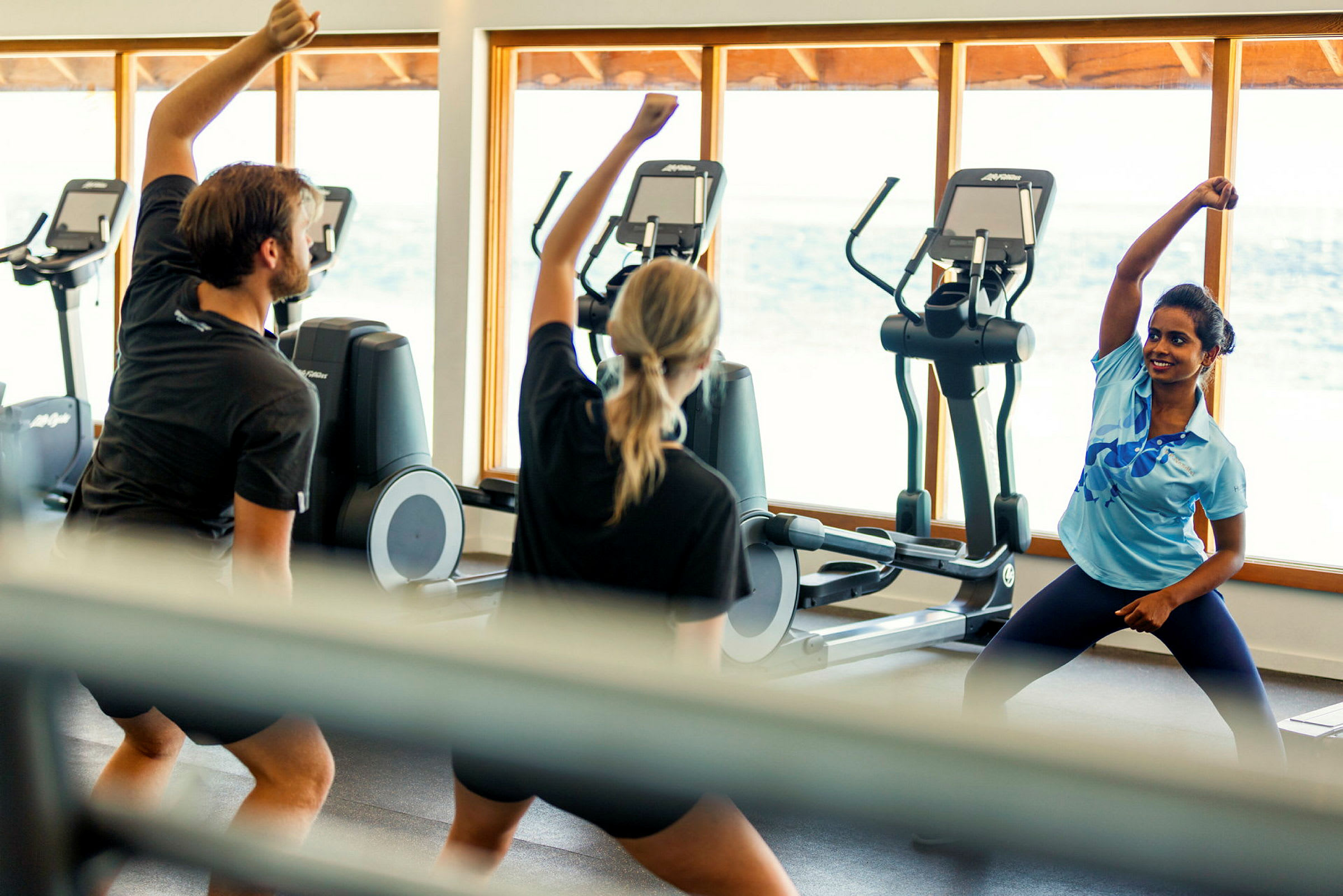 Personer udfører træningsøvelser i et fitnesscenter med konditionsmaskiner og store vinduer med udsigt over havet. (AI-genereret tekst)