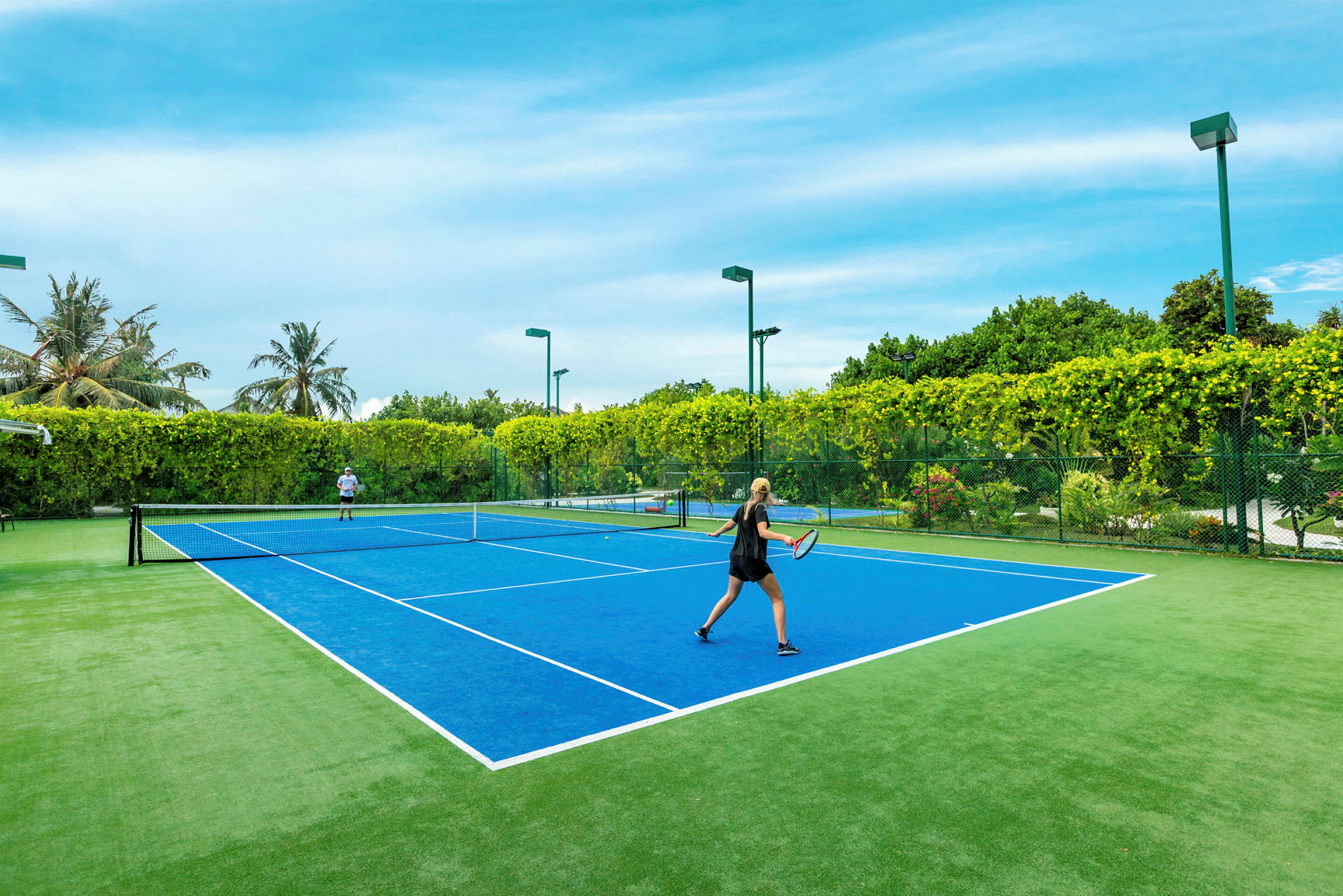 To personer spiller tennis på en blå tennisbane omgivet af grønt landskab. (AI-genereret tekst)