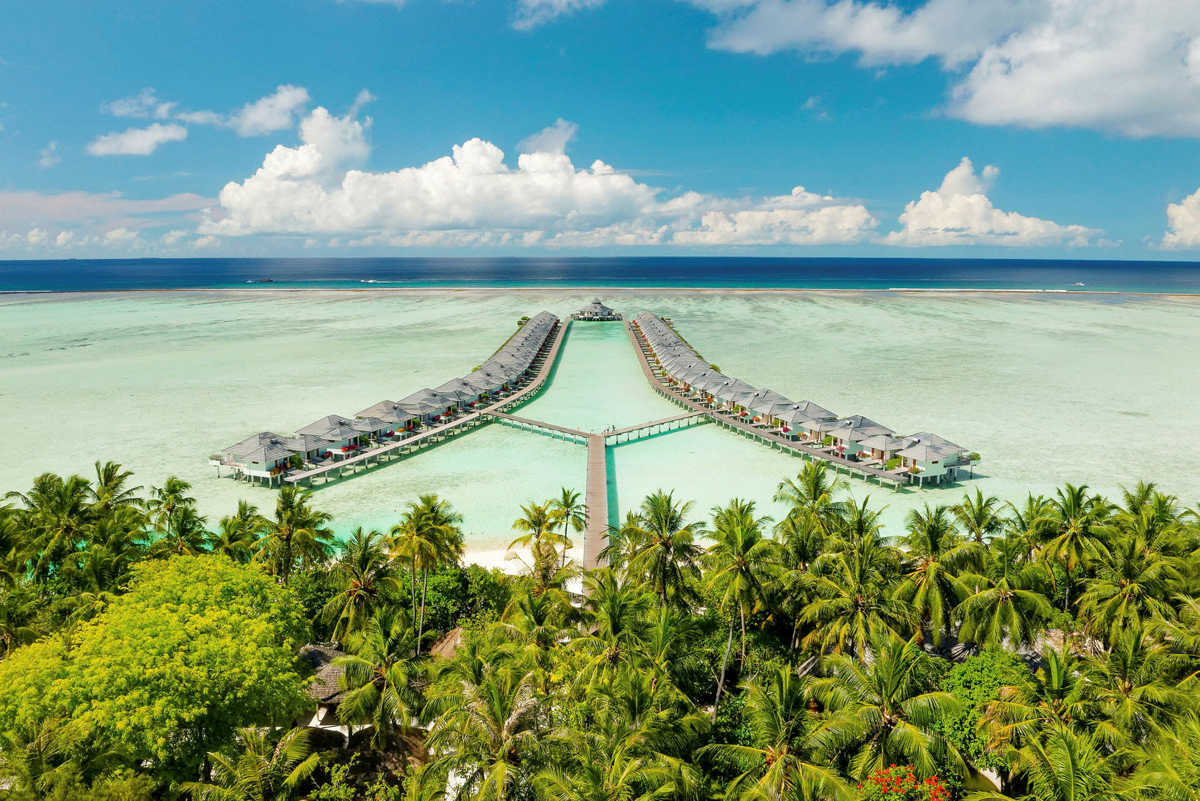 Billedet viser vandbungalower forbundet med gangbroer over en lagune, omgivet af palmer og omgivet af en blå himmel og hav. (AI-genereret tekst)