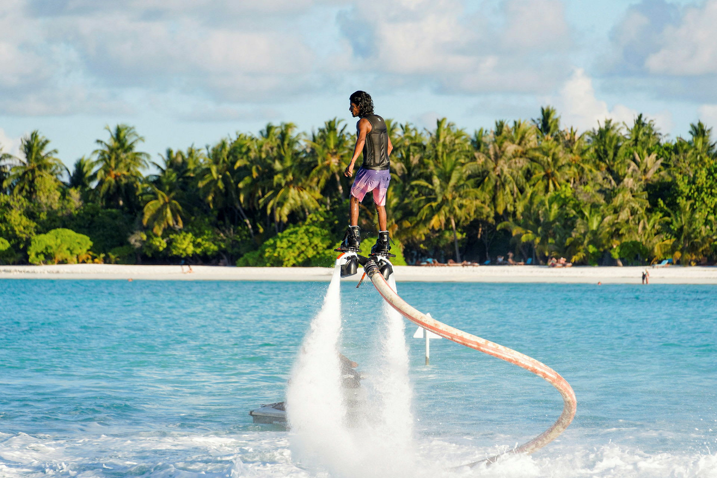 Person flyver på vandjet over havet med en hvid sandstrand og palmer i baggrunden. (AI-genereret tekst)