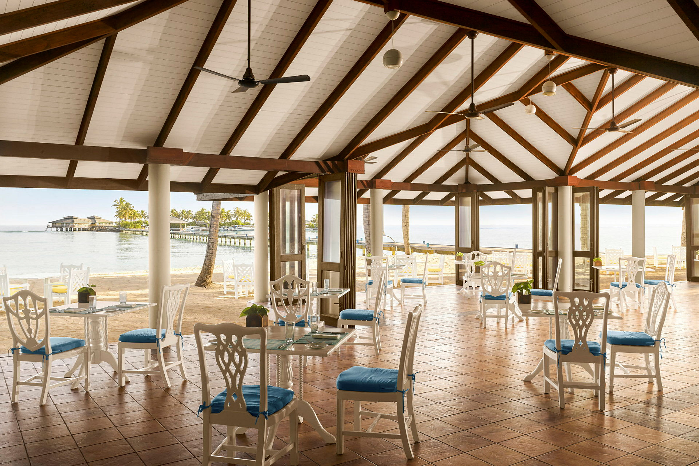 Billede af en åben restaurant med hvidmalede møbler og blå sædehynder, med udsigt over havet og en bro. (AI-genereret tekst)