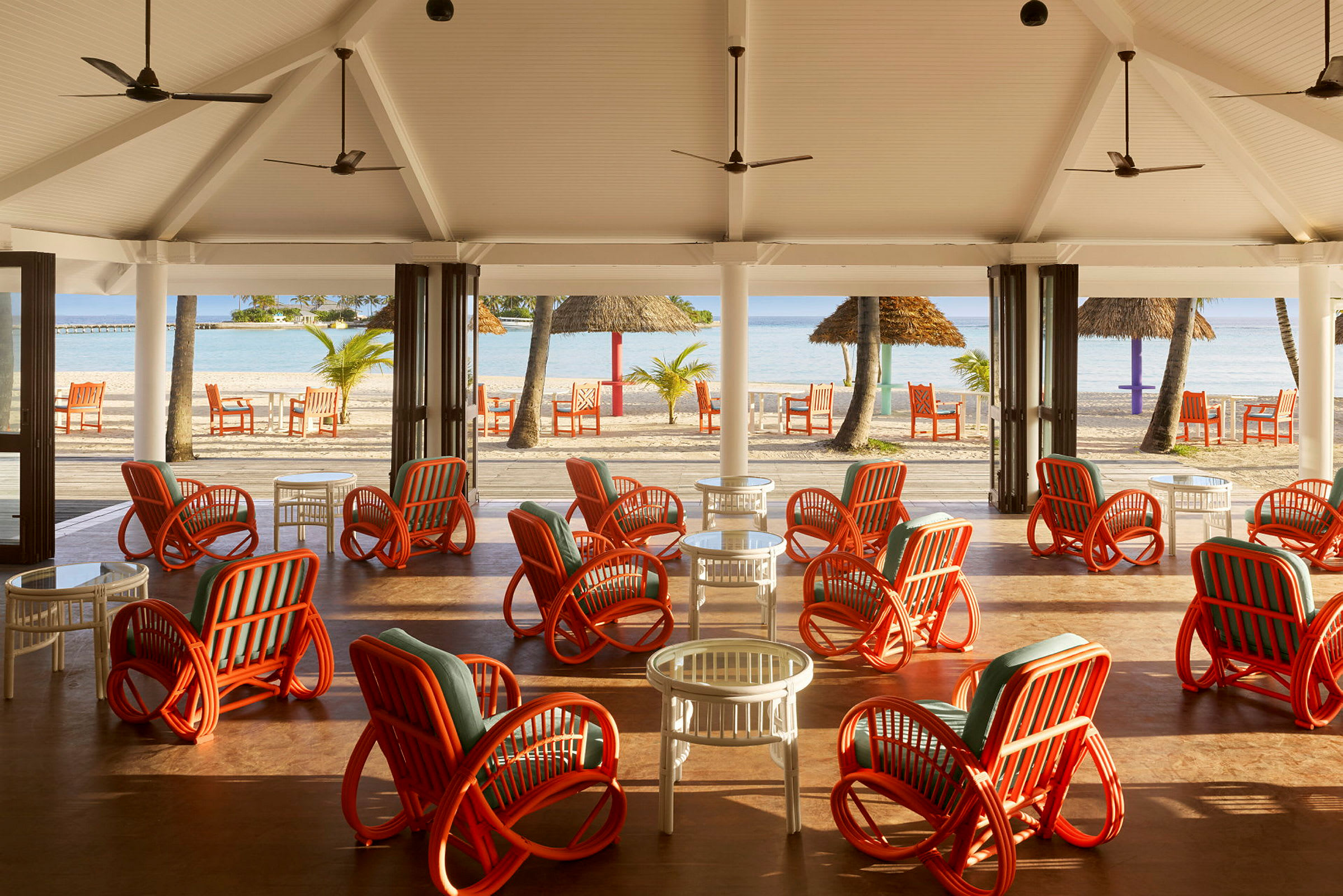 En åben lounge med farverige rattanstole og borde, med udsigt over stranden og havet. (AI-genereret tekst)