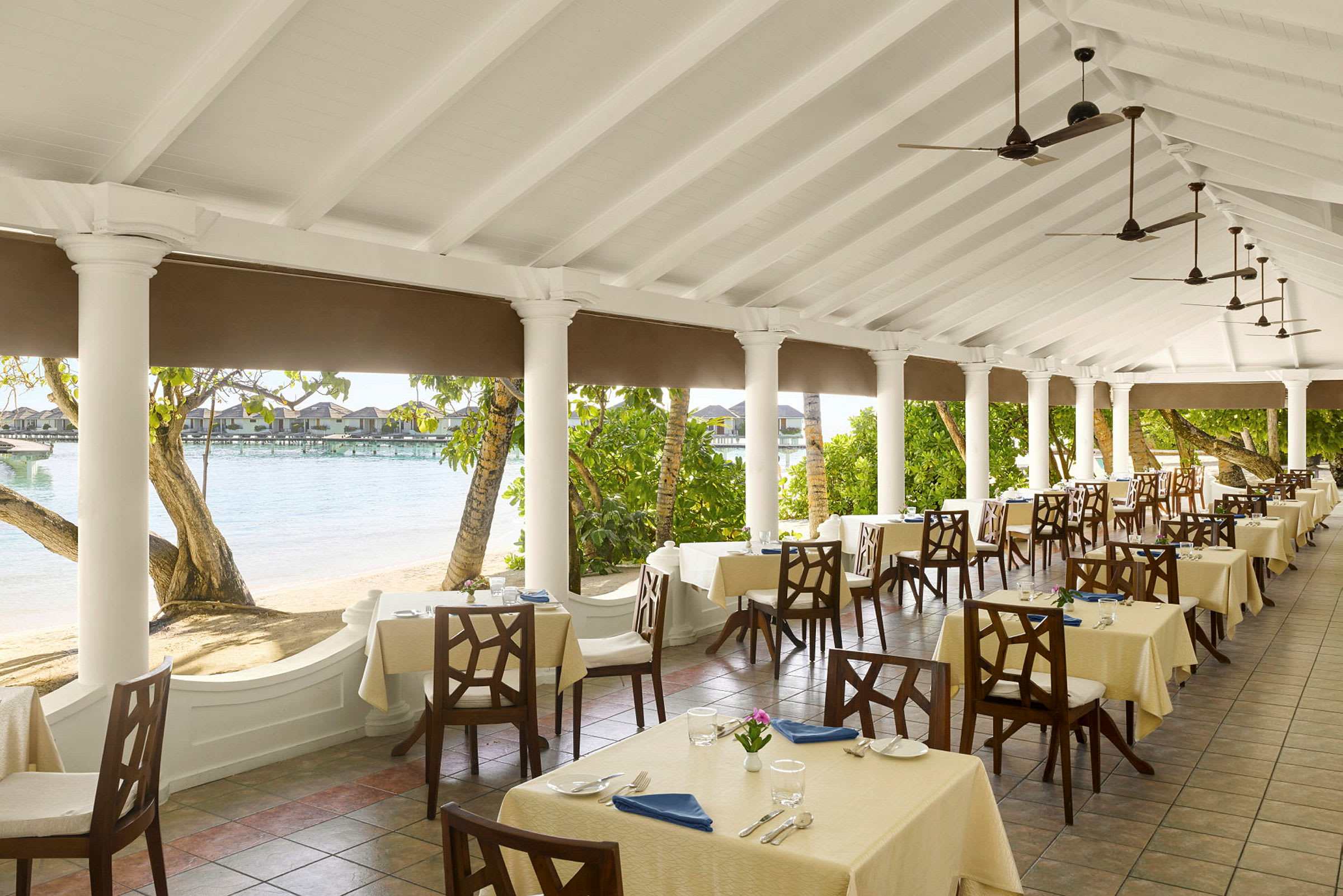 En restaurantterrasse med dækkede borde, omgivet af søjler og udsigt over stranden og vandet. (AI-genereret tekst)