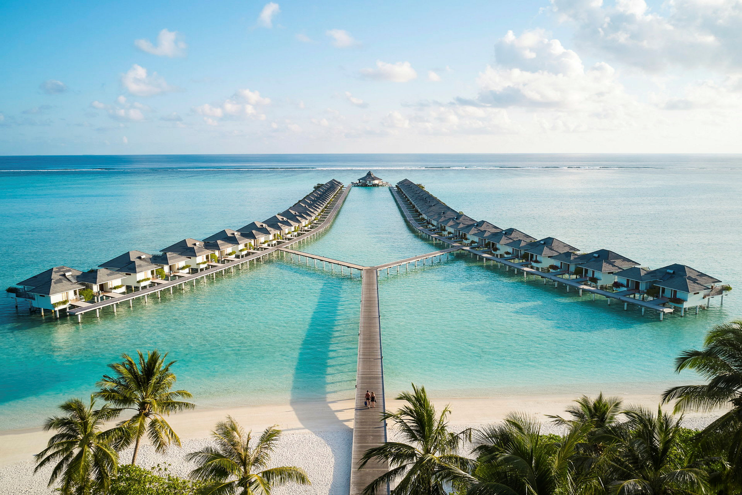 Hotelbungalower på pæle over klart blåt vand, med sandstrand og palmer i forgrunden. (AI-genereret tekst)