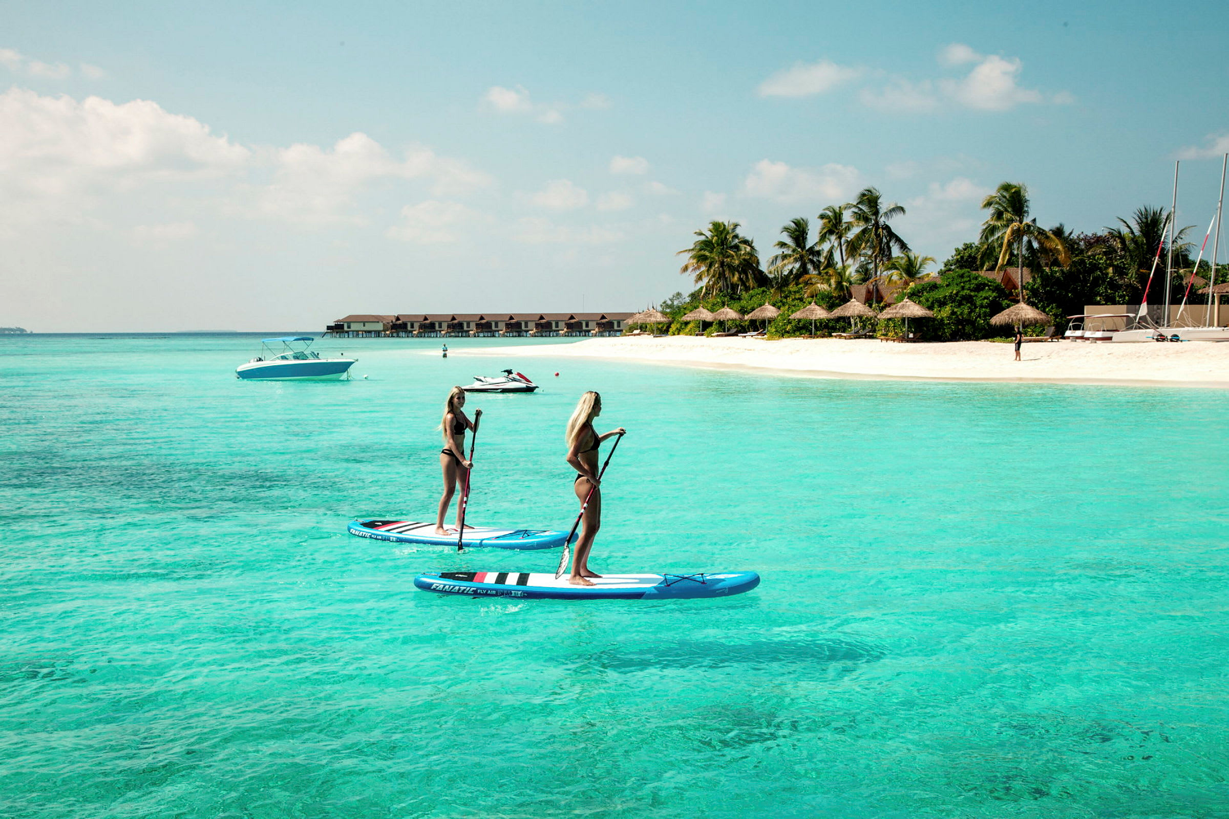 To personer står på paddleboards i klart turkis vand nær en sandstrand. (AI-genereret tekst)
