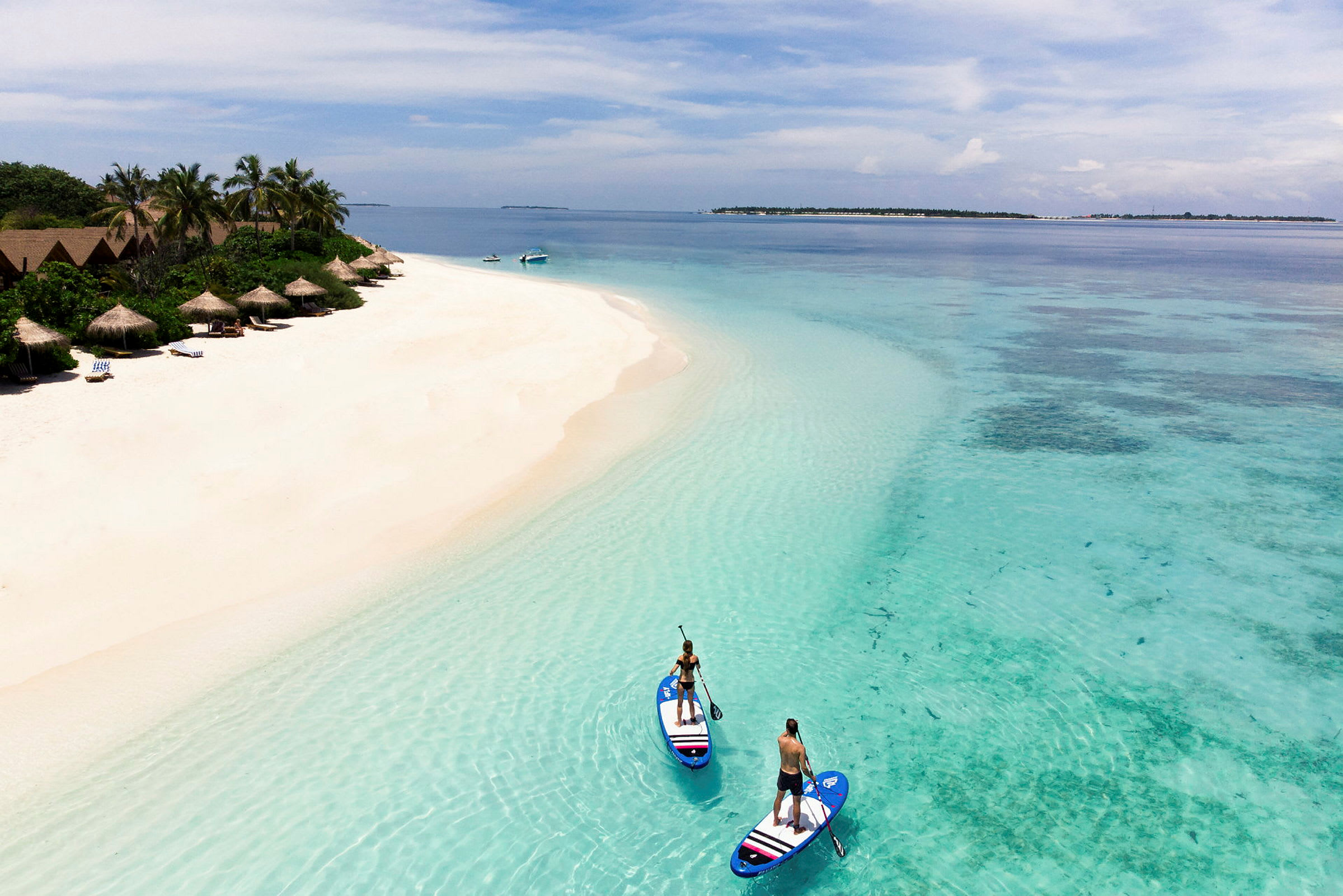 To personer står på paddleboards i klart turkisblåt vand ved en sandstrand med hytter og palmer. (AI-genereret tekst)