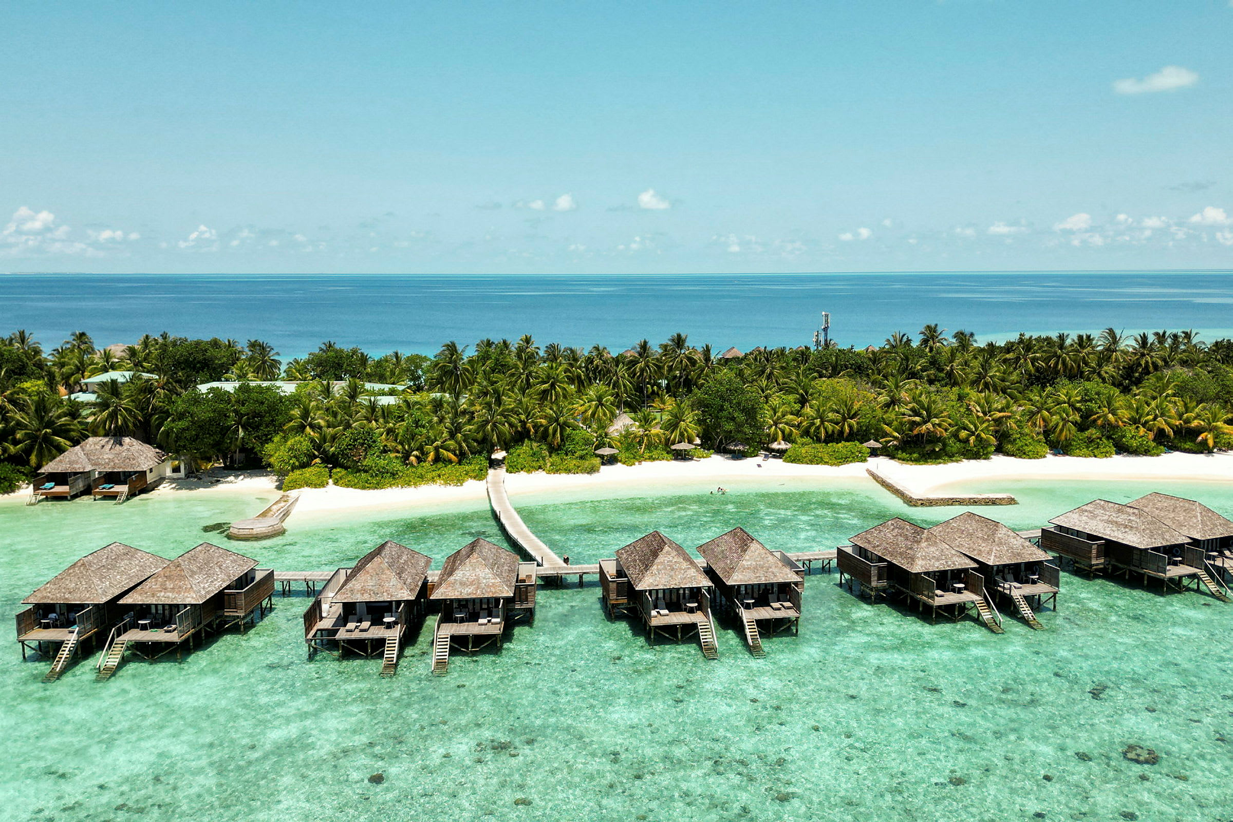Luksuriøse bungalower på pæle over klart turkis vand og en tropisk strand med palmer. (AI-genereret tekst)