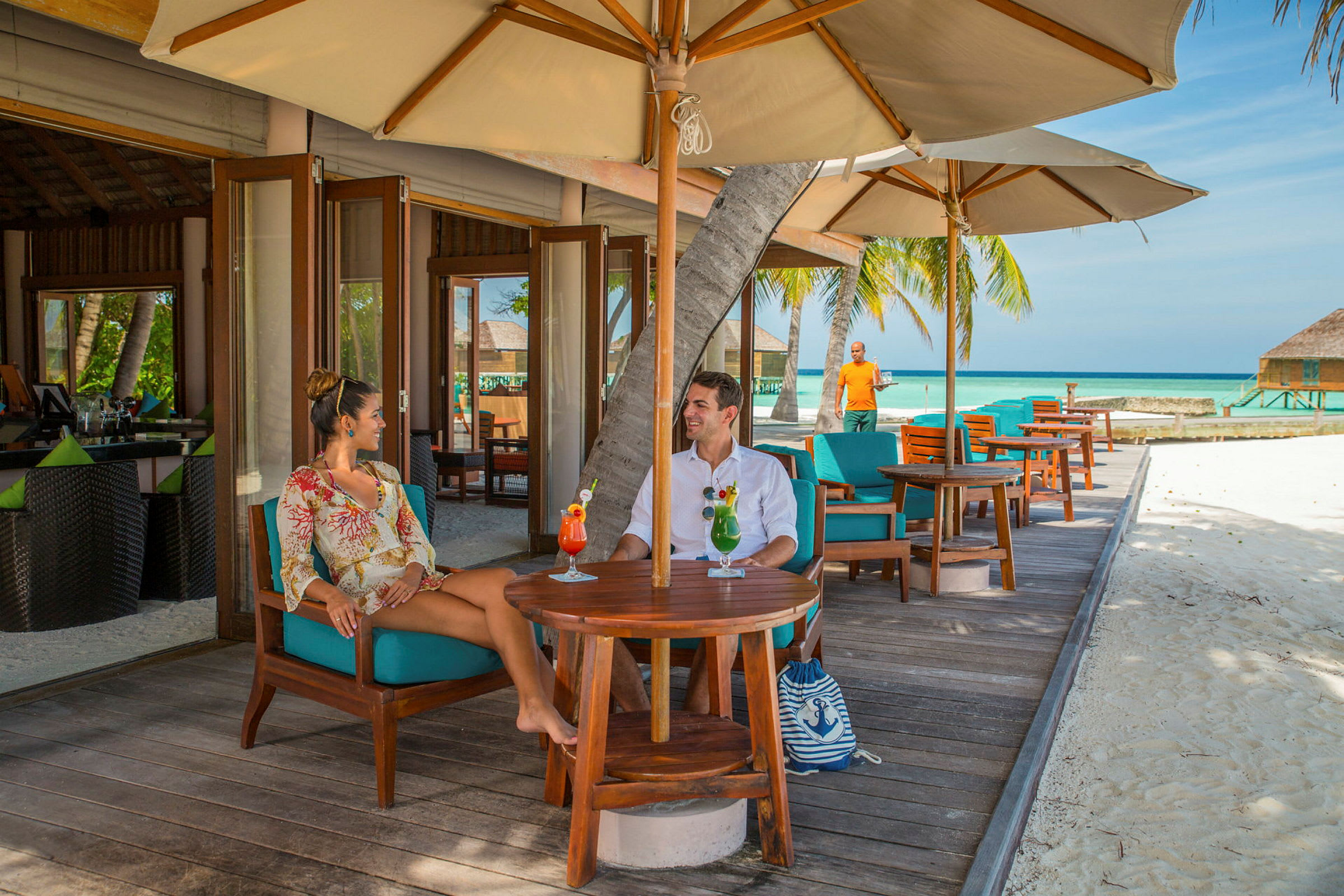 To personer sidder ved et bord på en udendørs restaurant med udsigt til sandstrand og hav. (AI-genereret tekst)