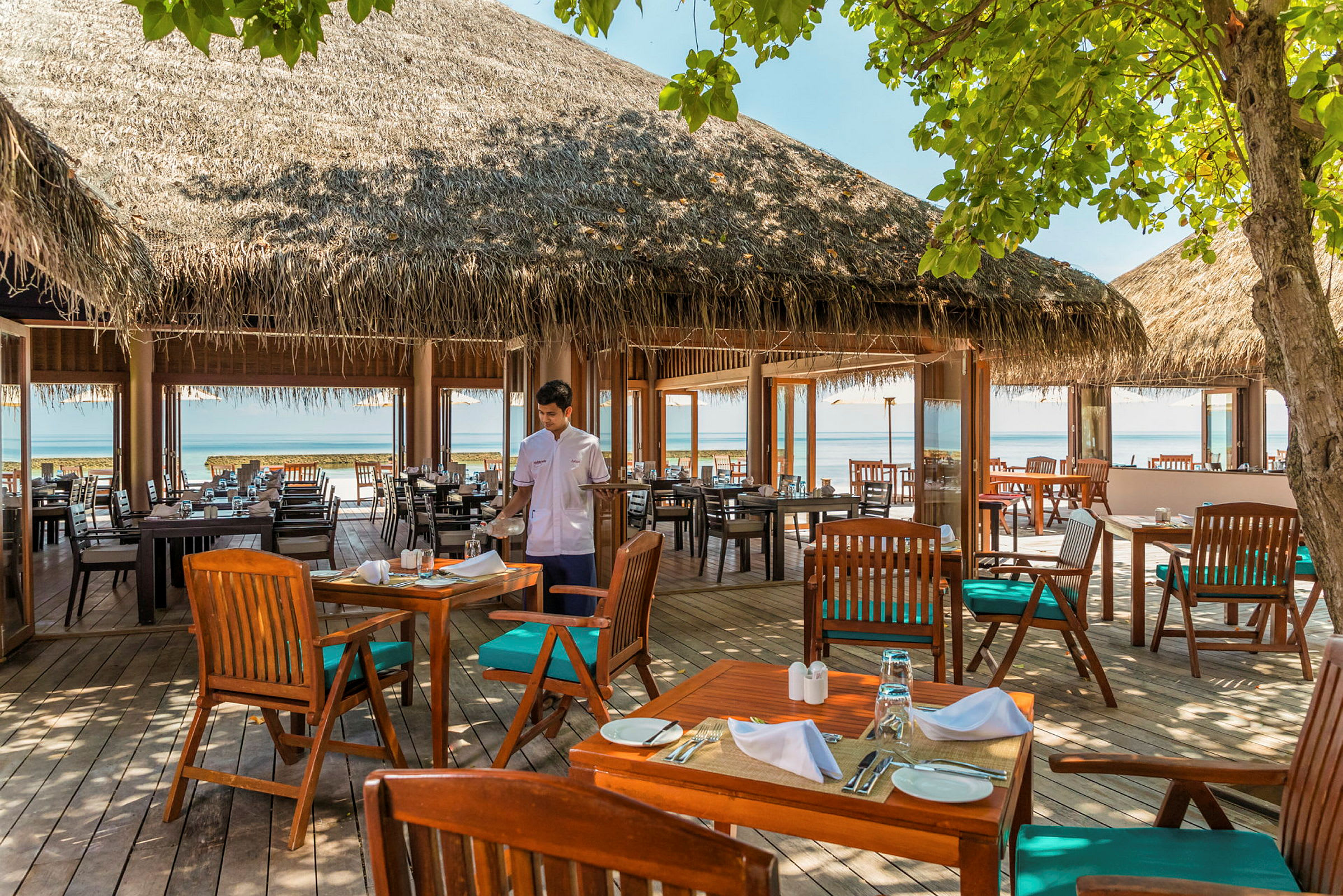 En udendørs restaurant med borde og stole af træ, stråtag og udsigt over havet. (AI-genereret tekst)