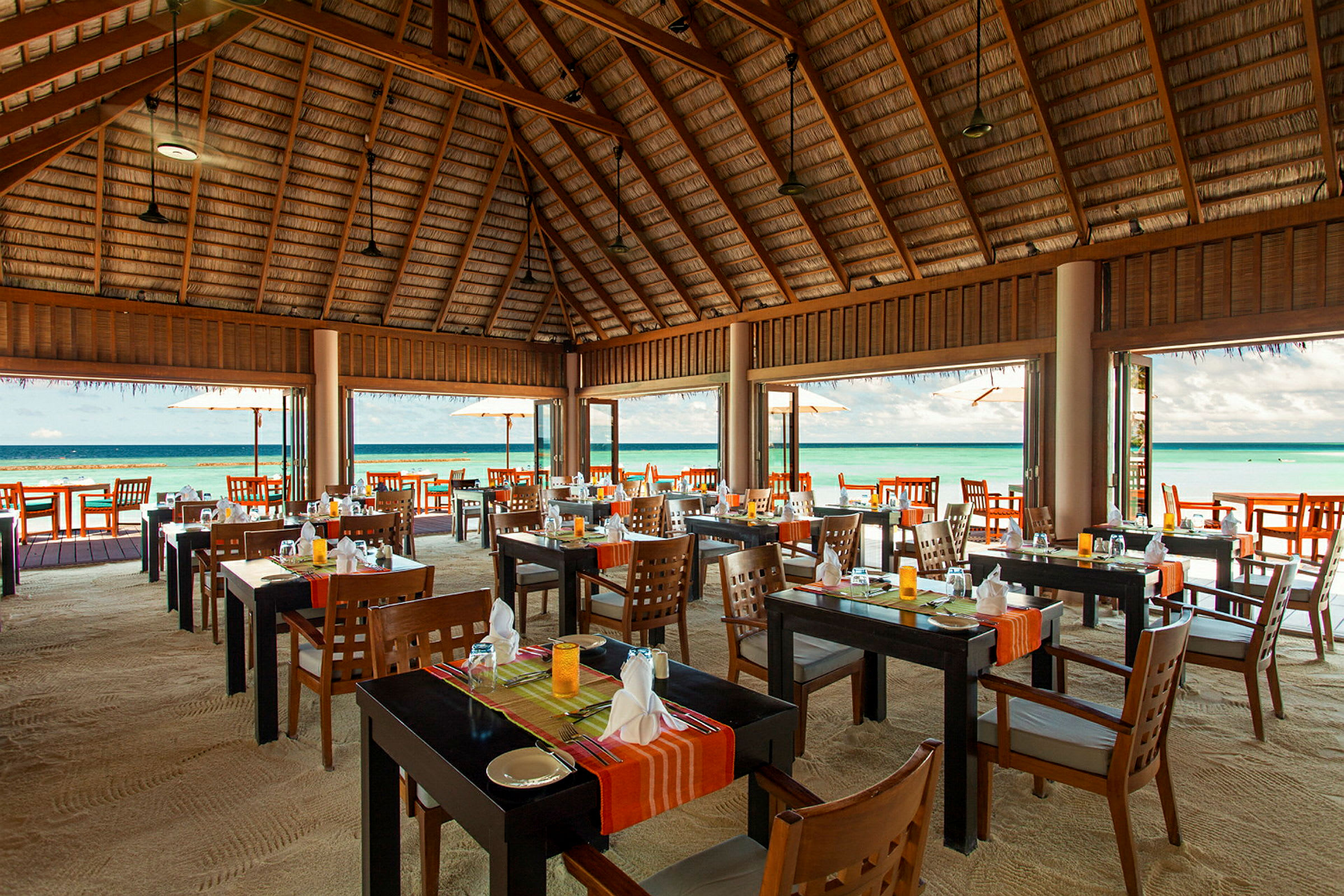 Restaurant med åbent trætak og borde på sand nær havet. (AI-genereret tekst)