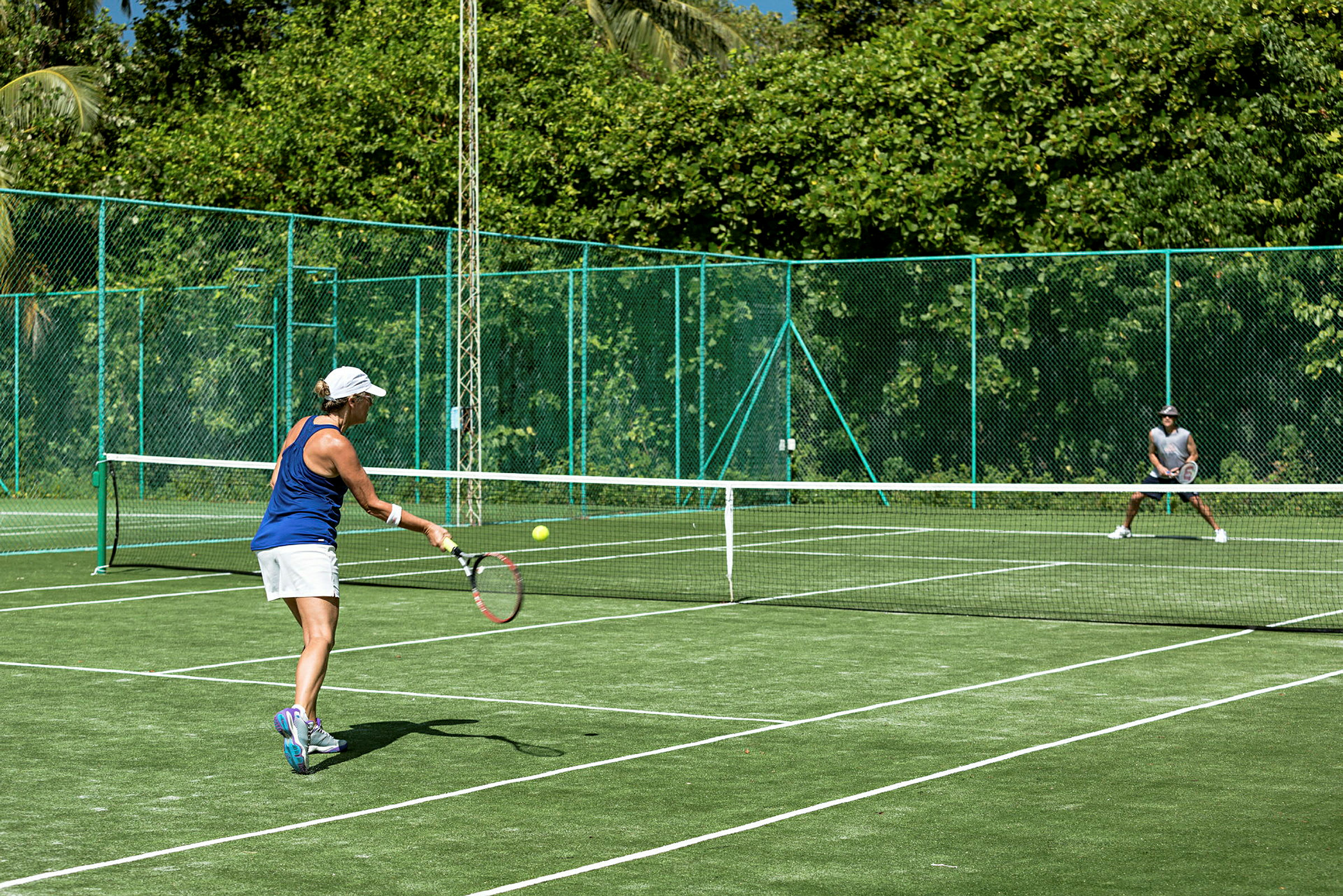 En person spiller tennis på en grøn tennisbane omgivet af frodigt område. (AI-genereret tekst)
