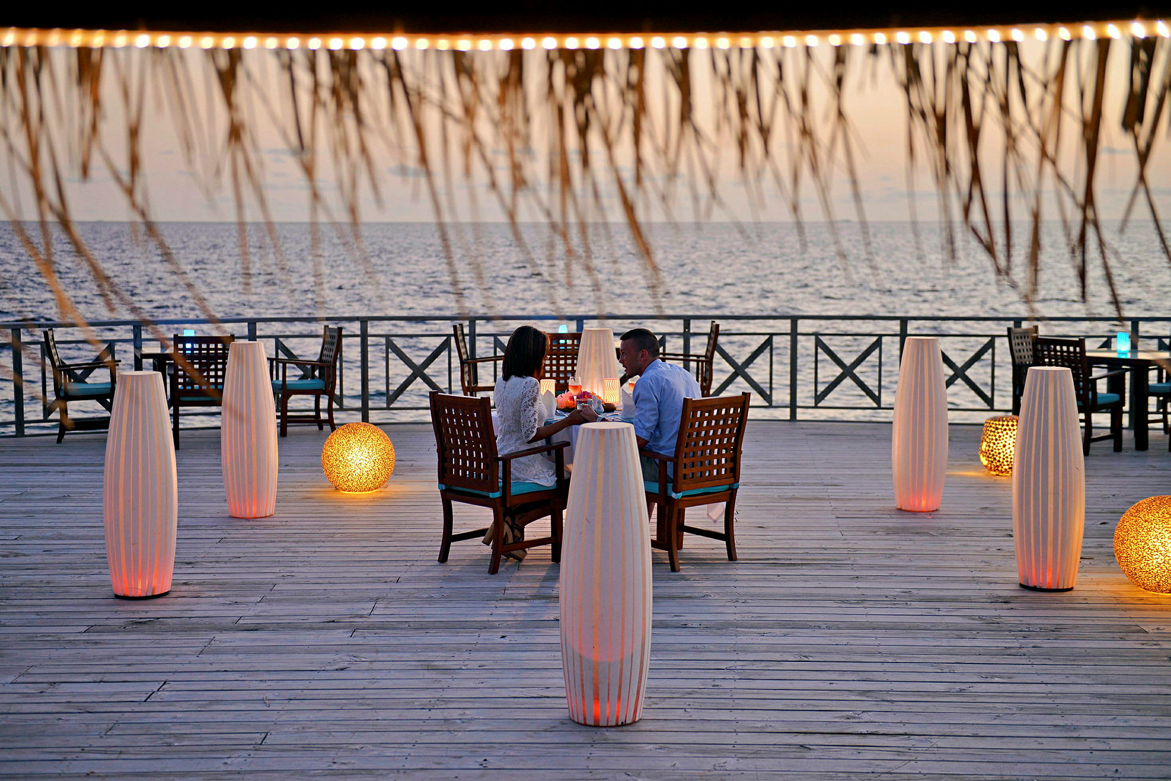 Personer sidder ved et bord på en træplatform ved havet, omgivet af dekorative lamper. (AI-genereret tekst)