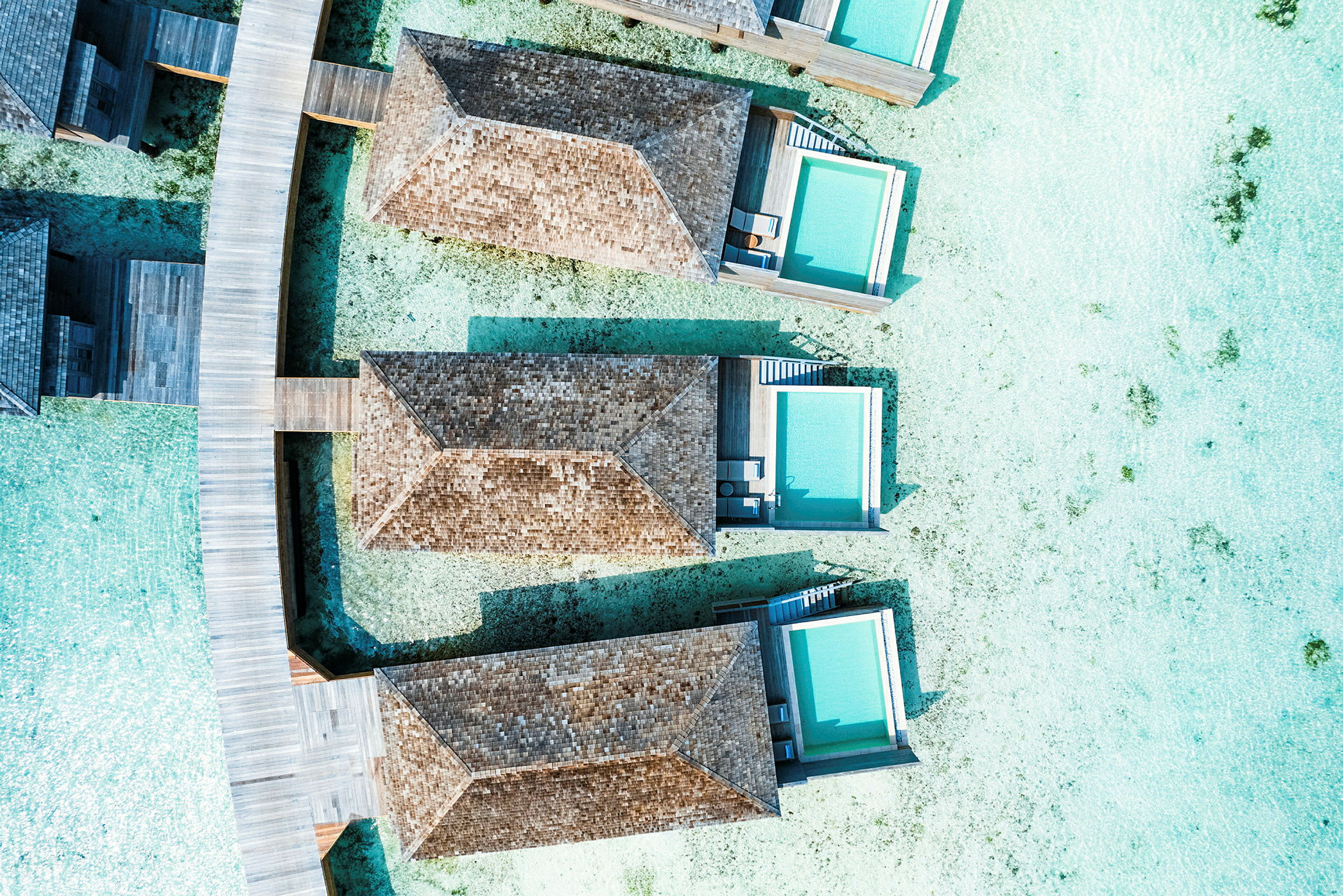 Luftfoto af strandvillaer med private pools bygget over klart vand, forbundet med træbroer. (AI-genereret tekst)