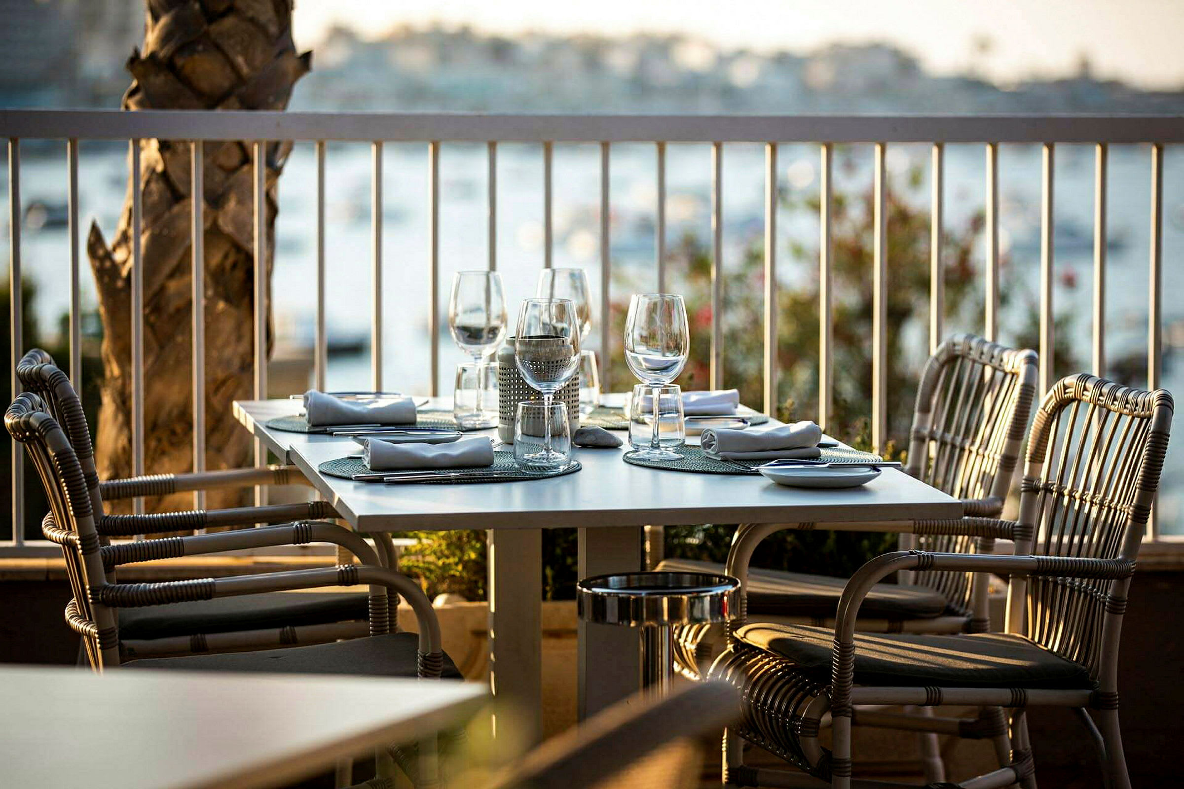 Et dækket bord med glas og servietter på en udendørs terrasse, omgivet af udsigt over vandet. (AI-genereret tekst)