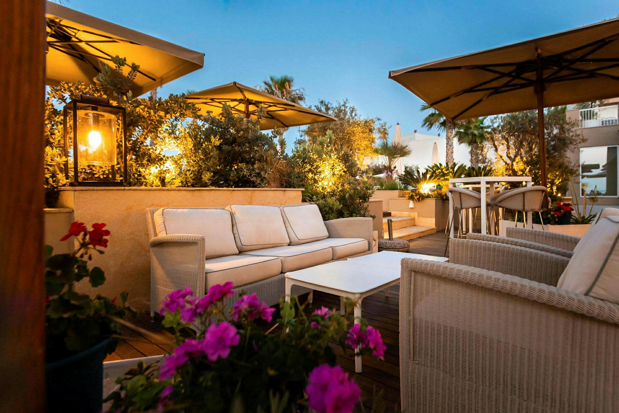 En terrasse med siddegrupper af kurvemøbler og parasoller, omgivet af planter og blomster. (AI-genereret tekst)