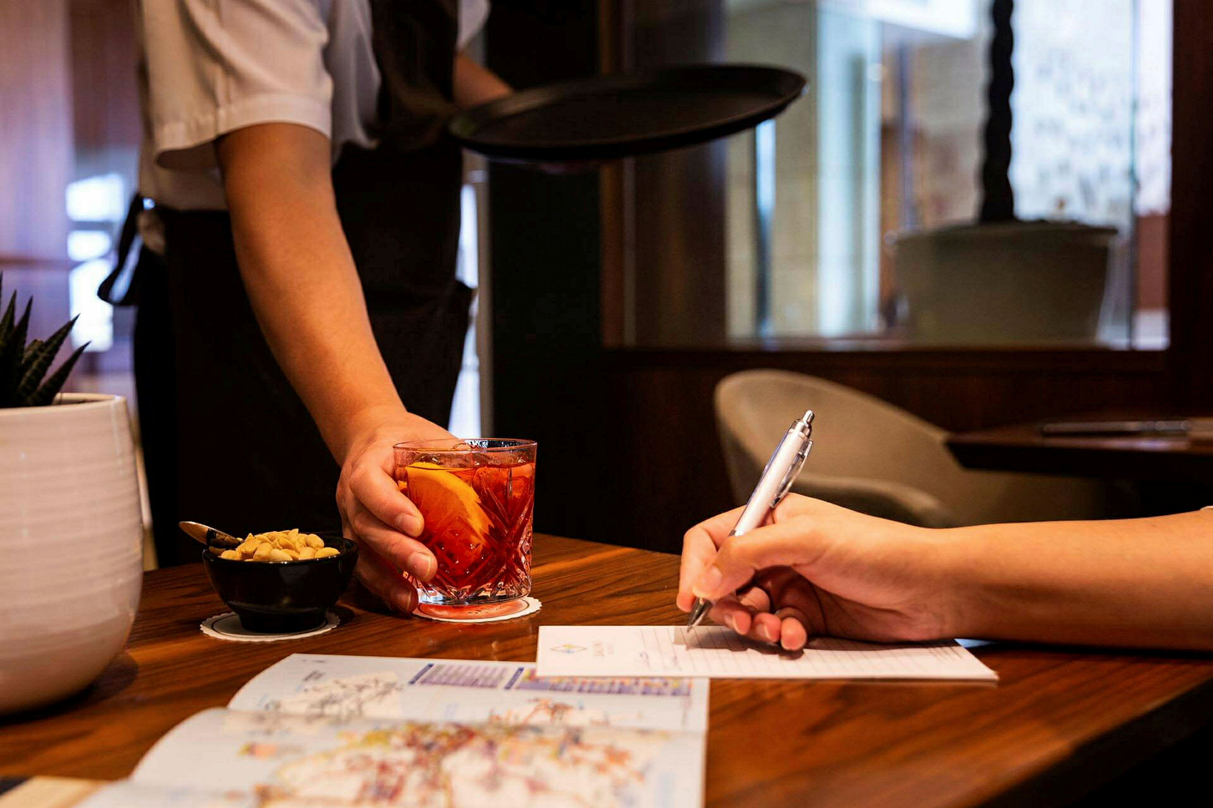 En tjener serverer en drink i et glas ved siden af en person, der skriver på et papir. (AI-genereret tekst)