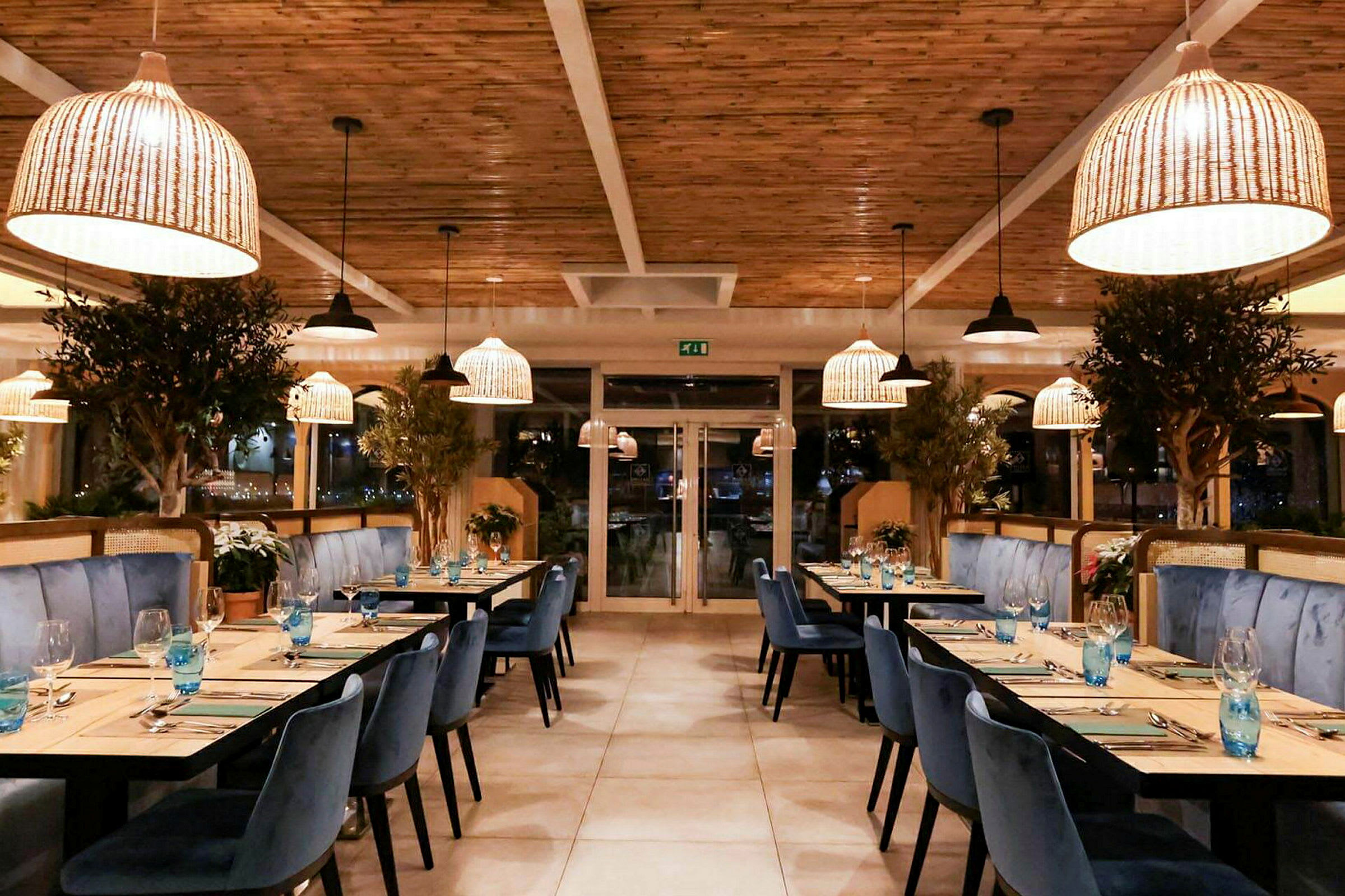 Et elegant restaurantinteriør med blå stole, træborde og store planteudsmykninger. Rottinglamper hænger fra loftet. (AI-genereret tekst)