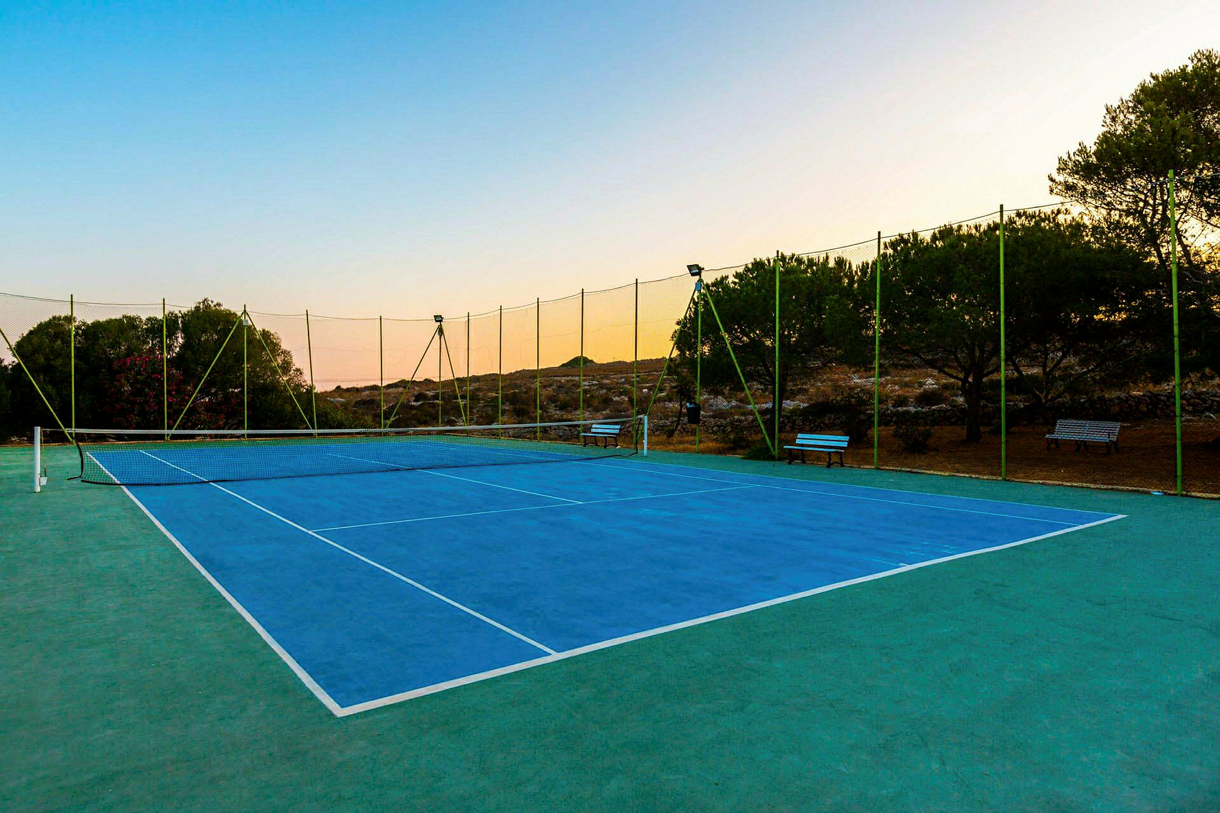 En tennisbane med blå belægning omgivet af grønt net og træer. (AI-genereret tekst)