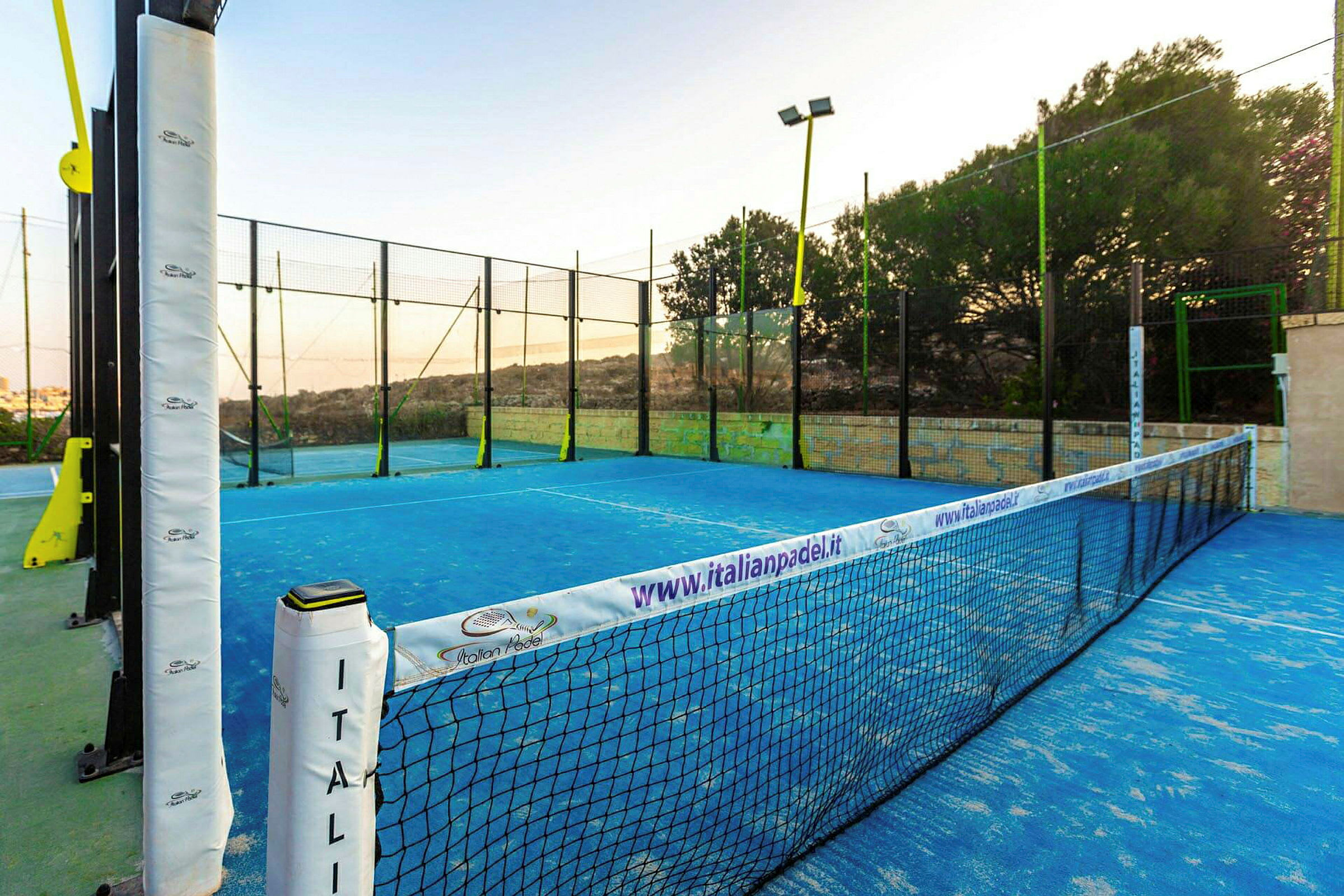 En padelbane med blå markeringer, omgivet af net og hegn i et udendørsmiljø. (AI-genereret tekst)