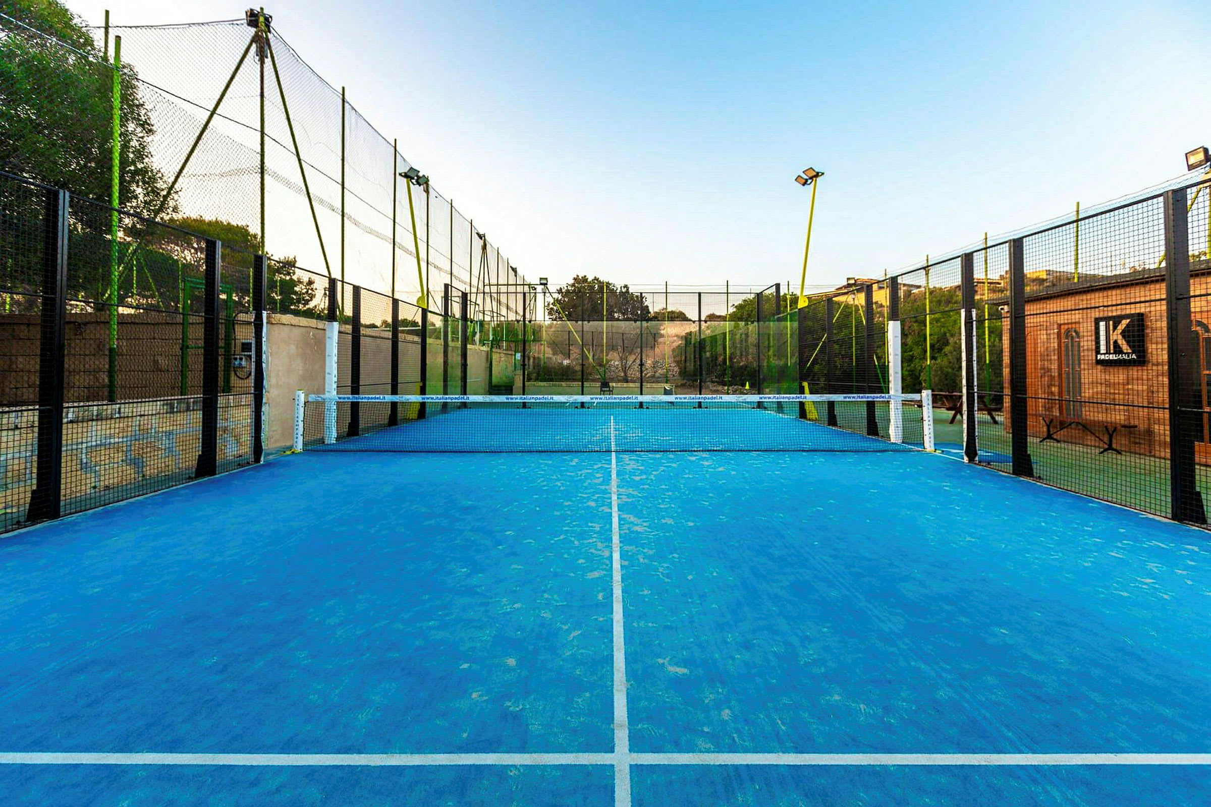 En padelbane med blå overflade og net i midten, omgivet af hegn. (AI-genereret tekst)