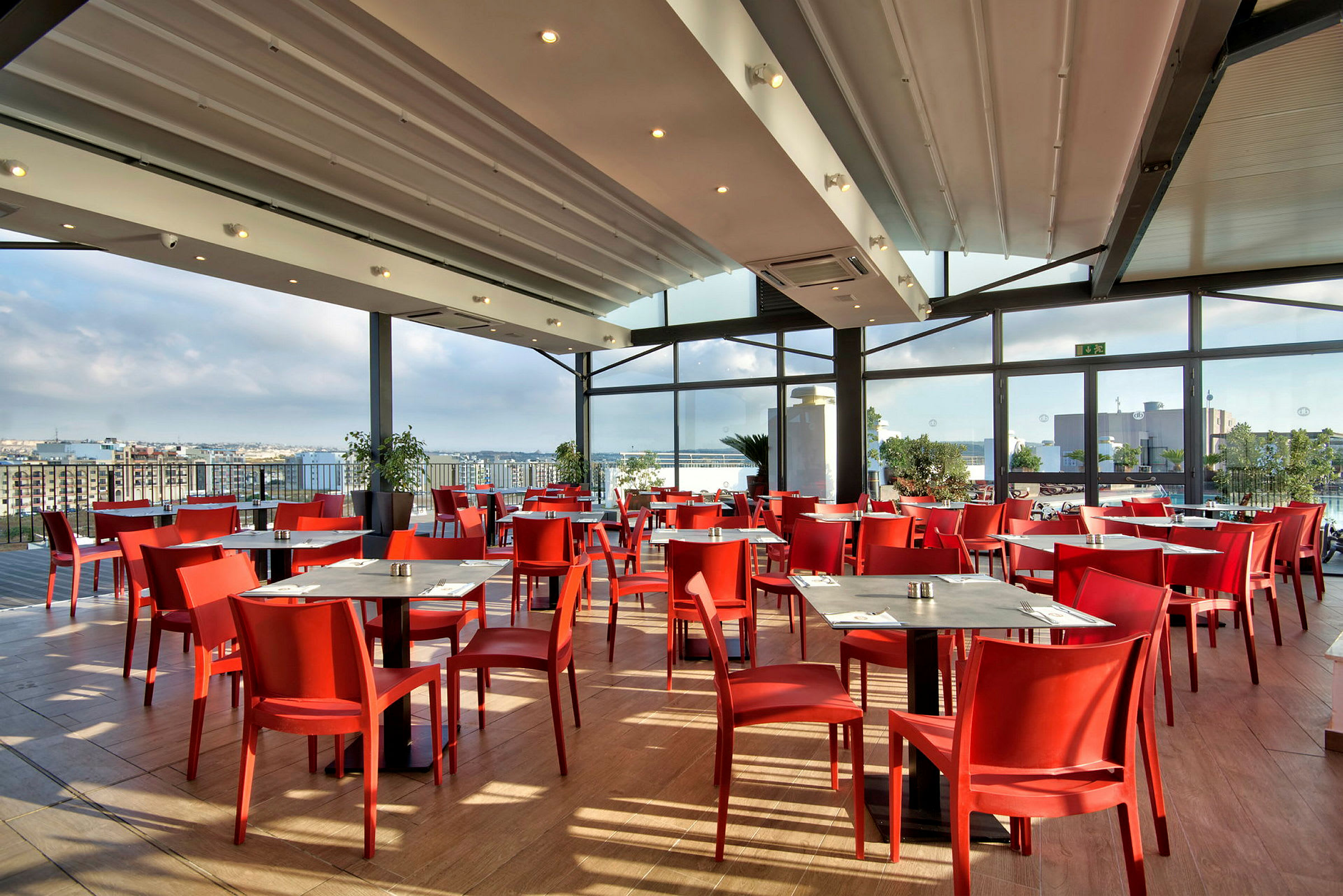 En lys restaurant med røde stole, træborde og store vinduer med udsigt over en by. (AI-genereret tekst)