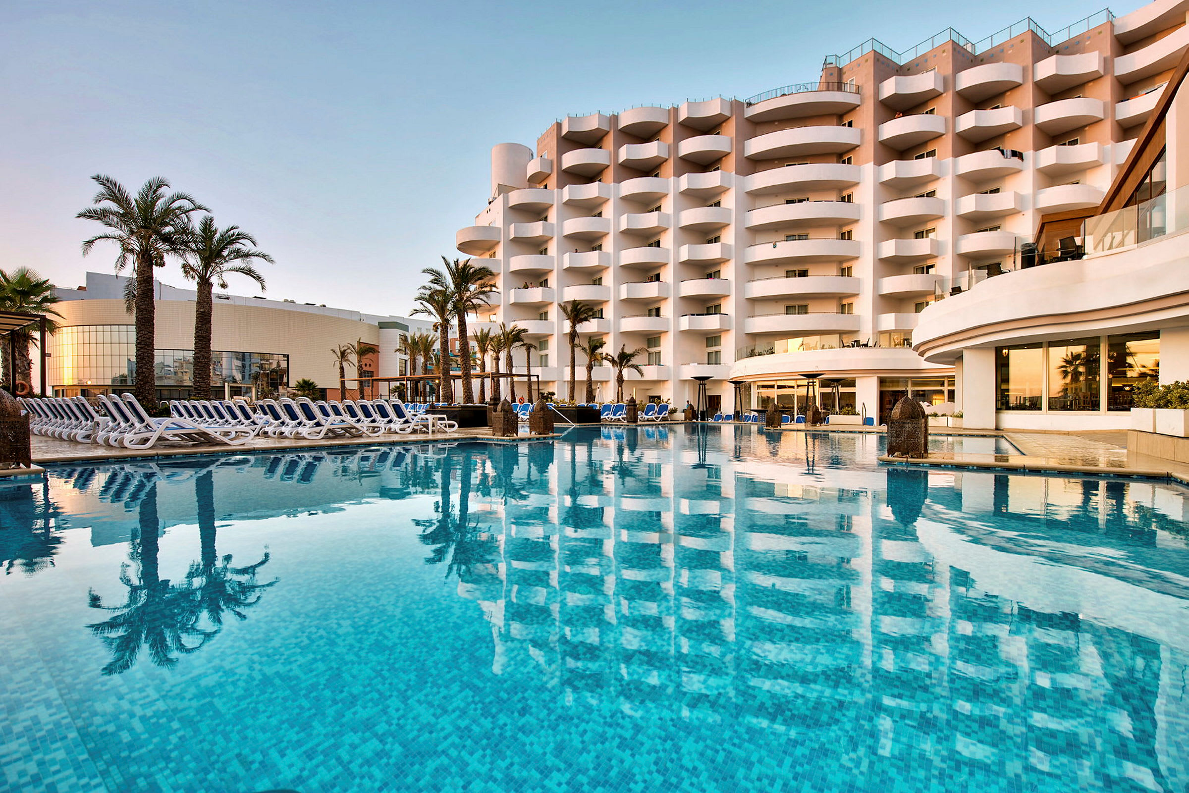 Hotelbygning ved en udendørs pool med stole og palmer. (AI-genereret tekst)